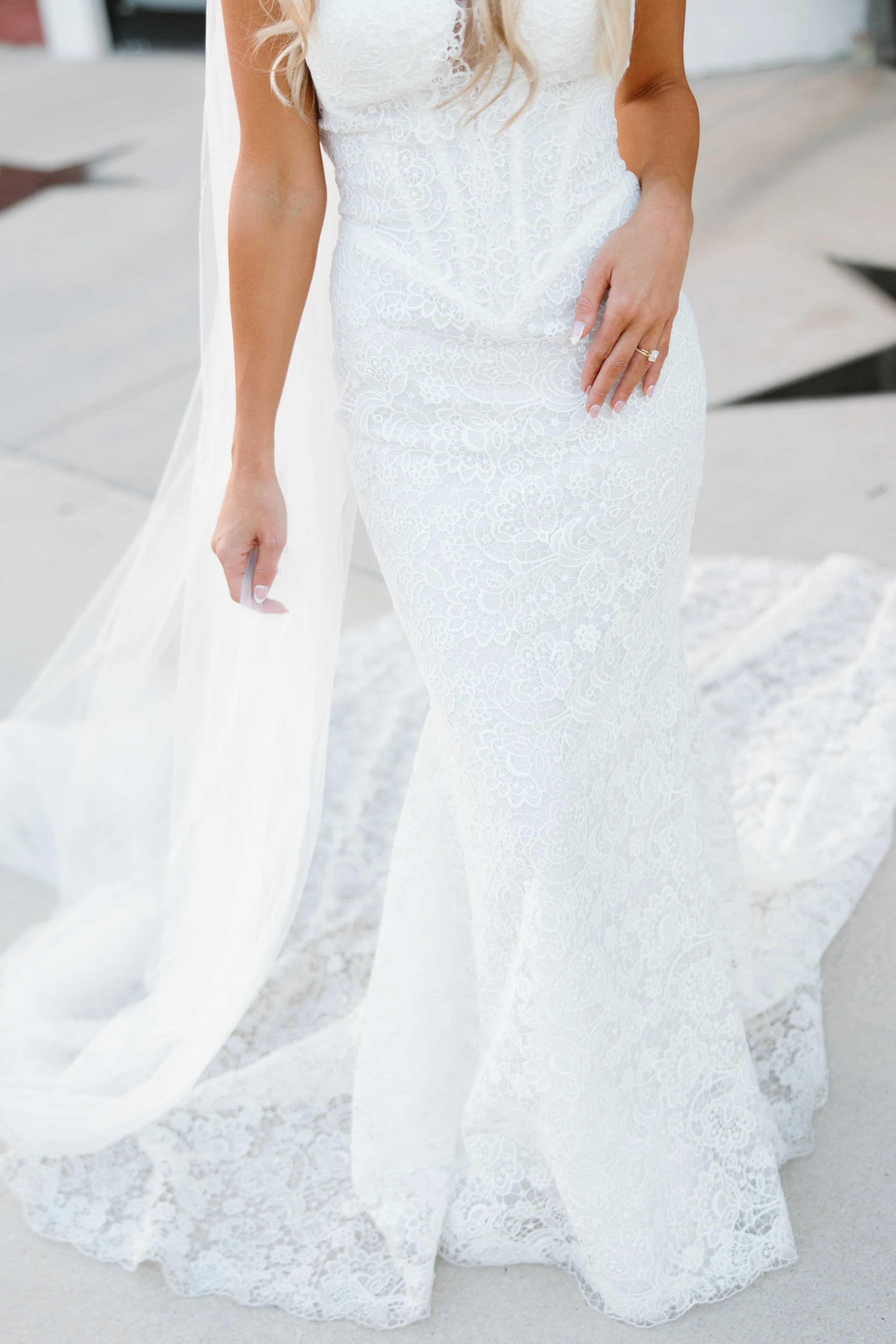 Close-up of a woman wearing a white lace wedding dress, holding her hand with an engagement ring, with a long flowing train, standing on a concrete surface.