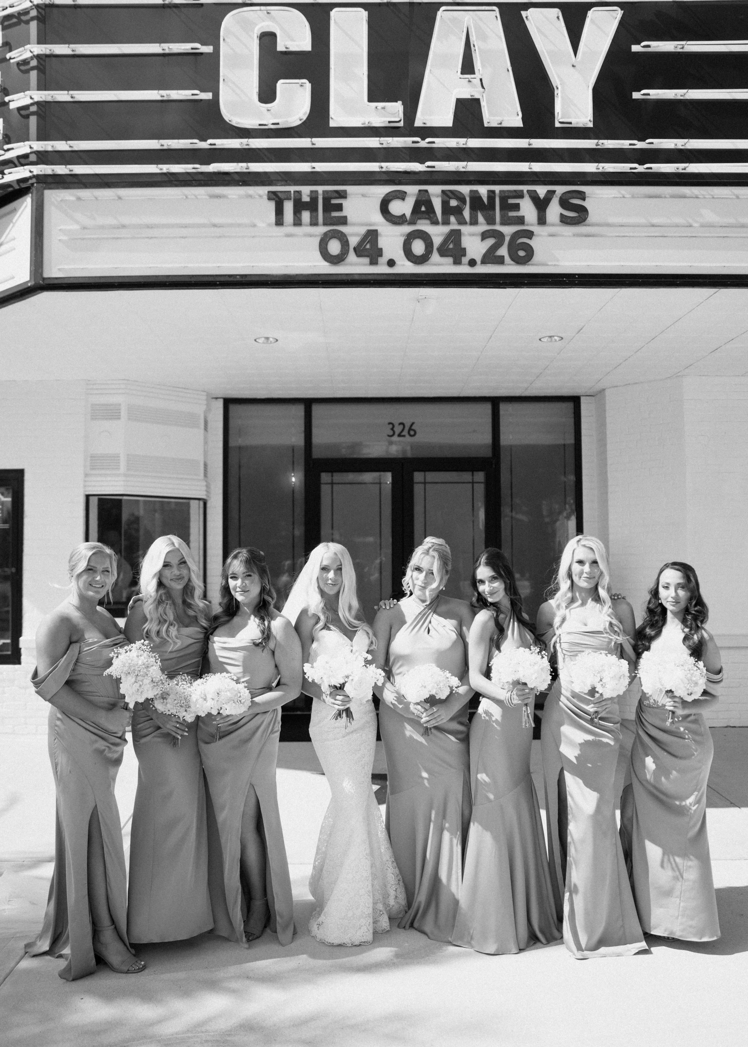 A group of nine women in formal dresses and holding bouquets, standing in front of a theater marquee that reads 'The Carneys 04.04.26'.
