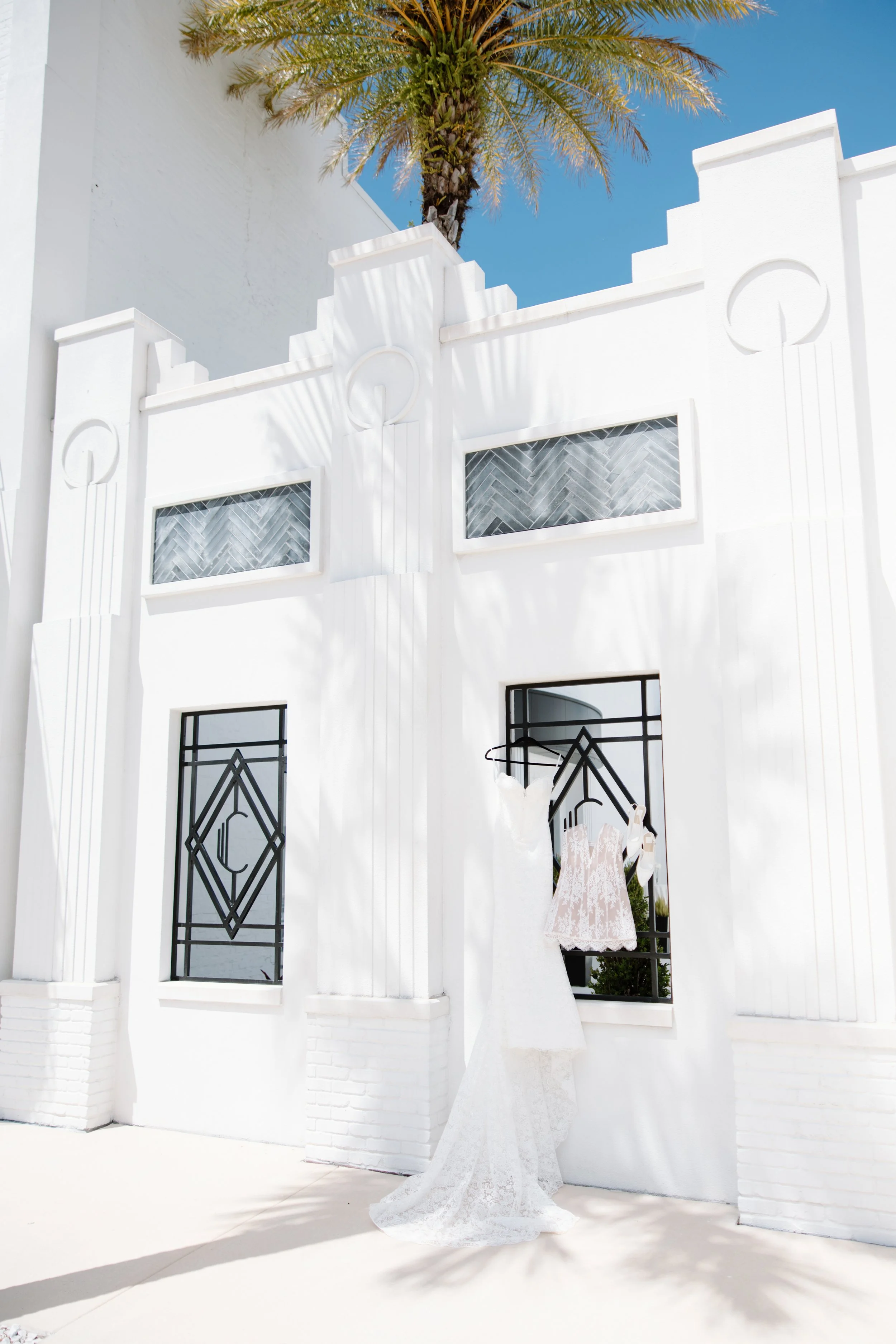 A white building with Art Deco design elements, featuring black decorative window grilles, a palm tree behind the building, and a wedding dress displayed on a mannequin with sunlight casting shadows.