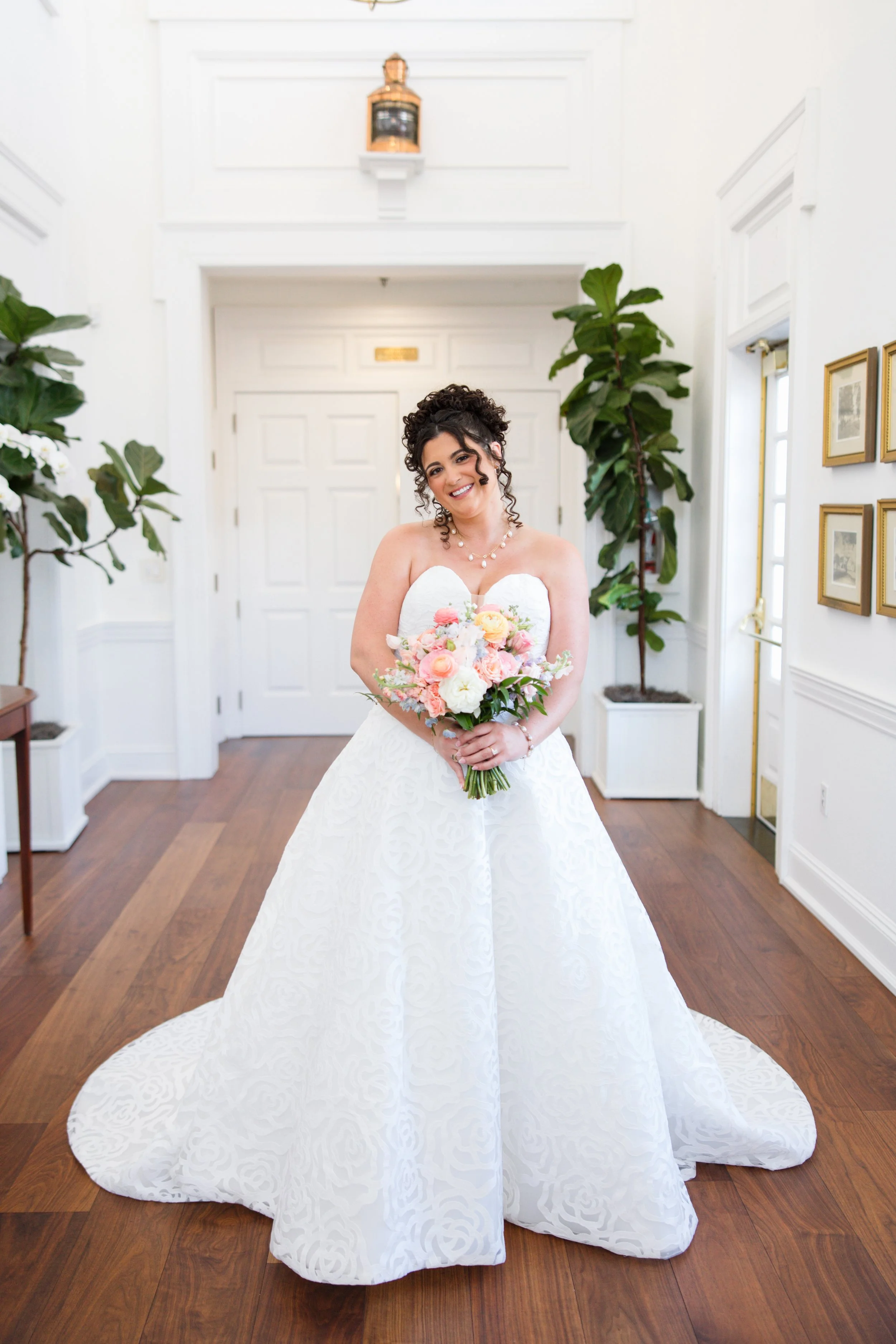 A woman wearing a strapless white wedding dress and holding a bouquet of pink, white, and peach roses stands indoors in front of a white door, with large green plants on either side and framed pictures on the wall to the right.