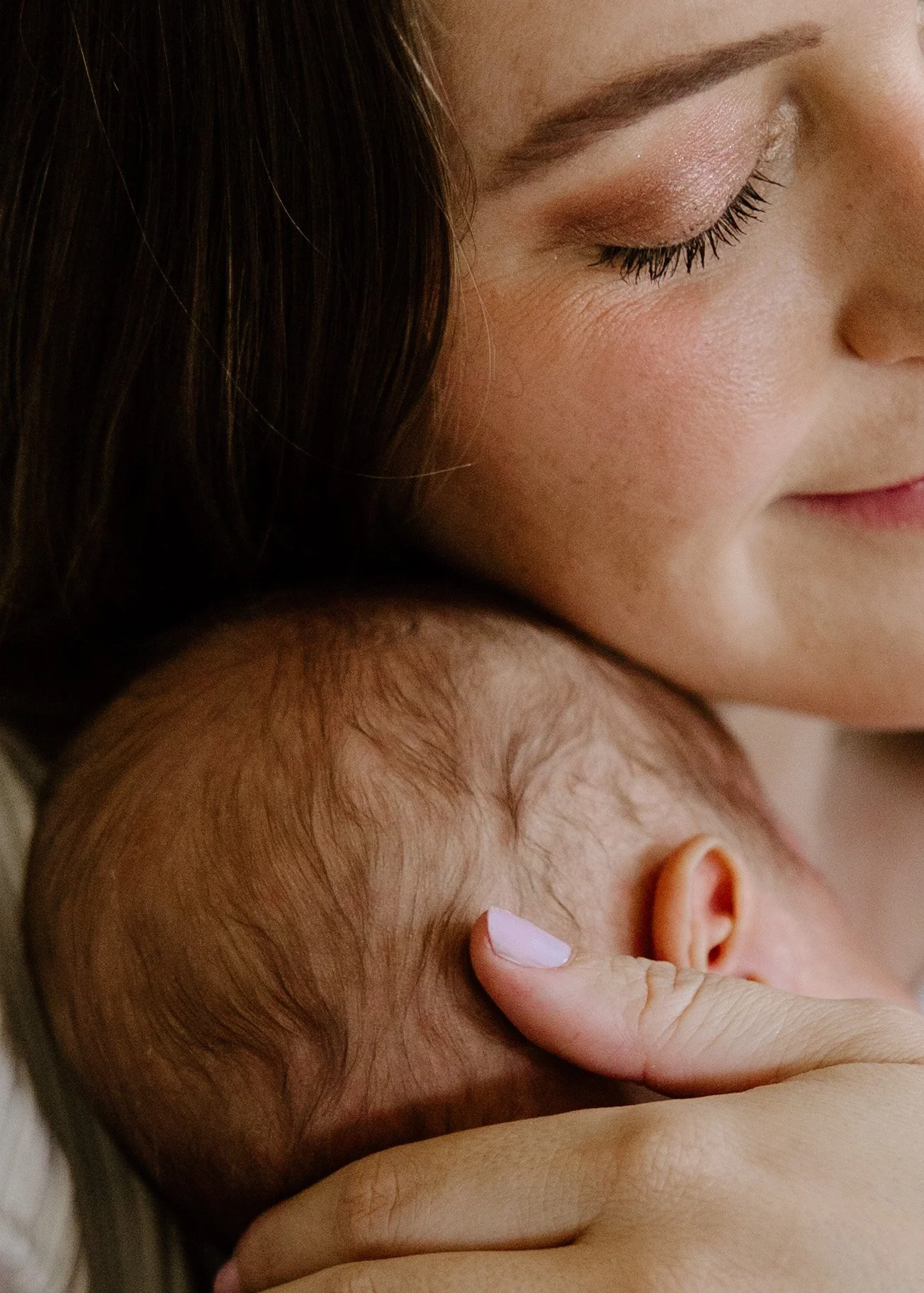 A woman with brown hair and makeup gently holding a newborn baby with light hair and closed eyes.