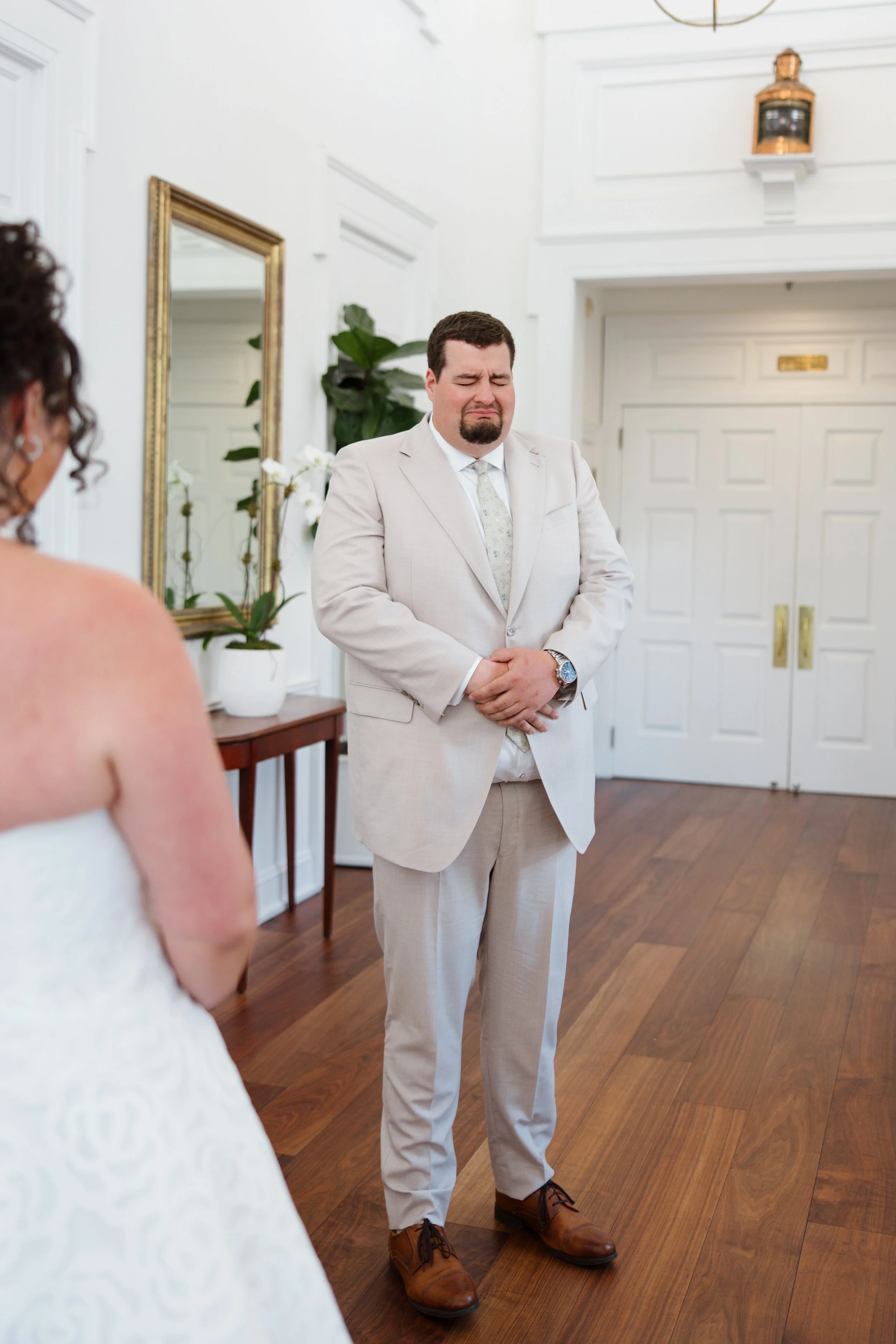 A man in a beige suit with a tearful or emotional expression stands with his hands clasped in front of him. A woman in a white dress is partially visible in the foreground, and the setting appears to be a formal event or ceremony, possibly a wedding,