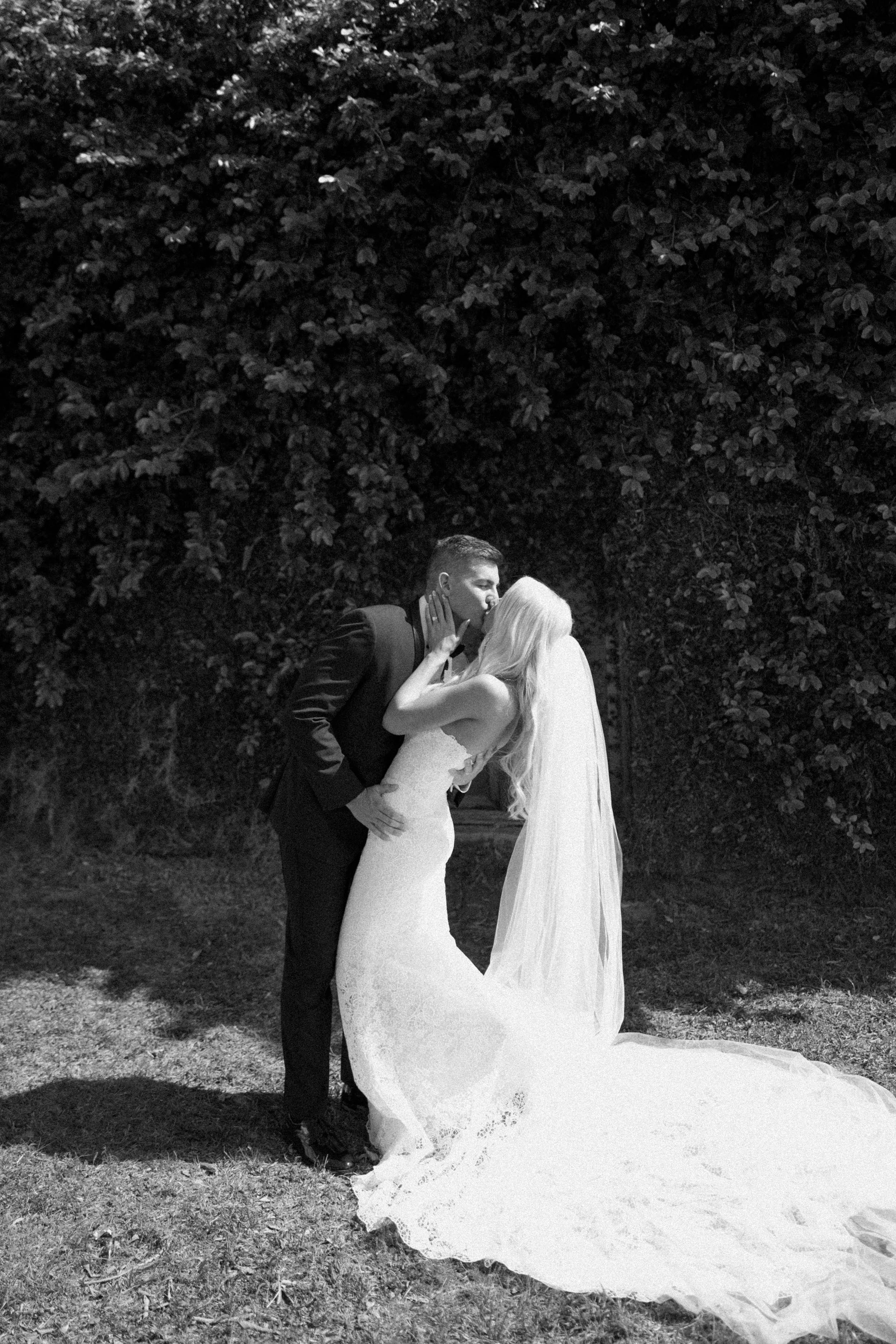 A couple in wedding attire kissing outdoors in front of a large bush. The groom is in a tuxedo, and the bride is in a lace wedding gown with a long train.