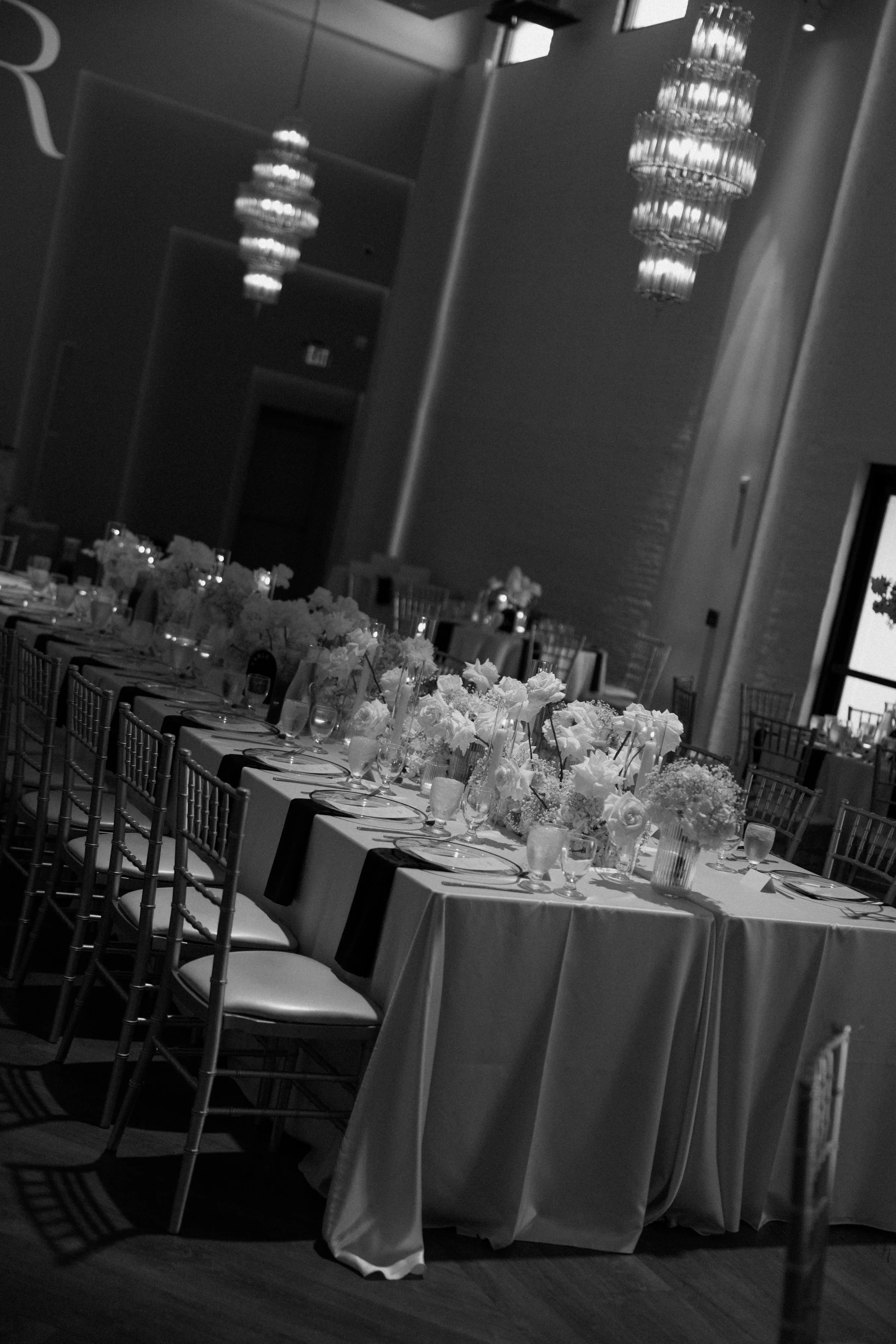 A banquet hall decorated for a formal event with a long table adorned with floral centerpieces and set with glassware and plates, elegant chandeliers hanging from the ceiling, and chairs arranged along the sides of the table.
