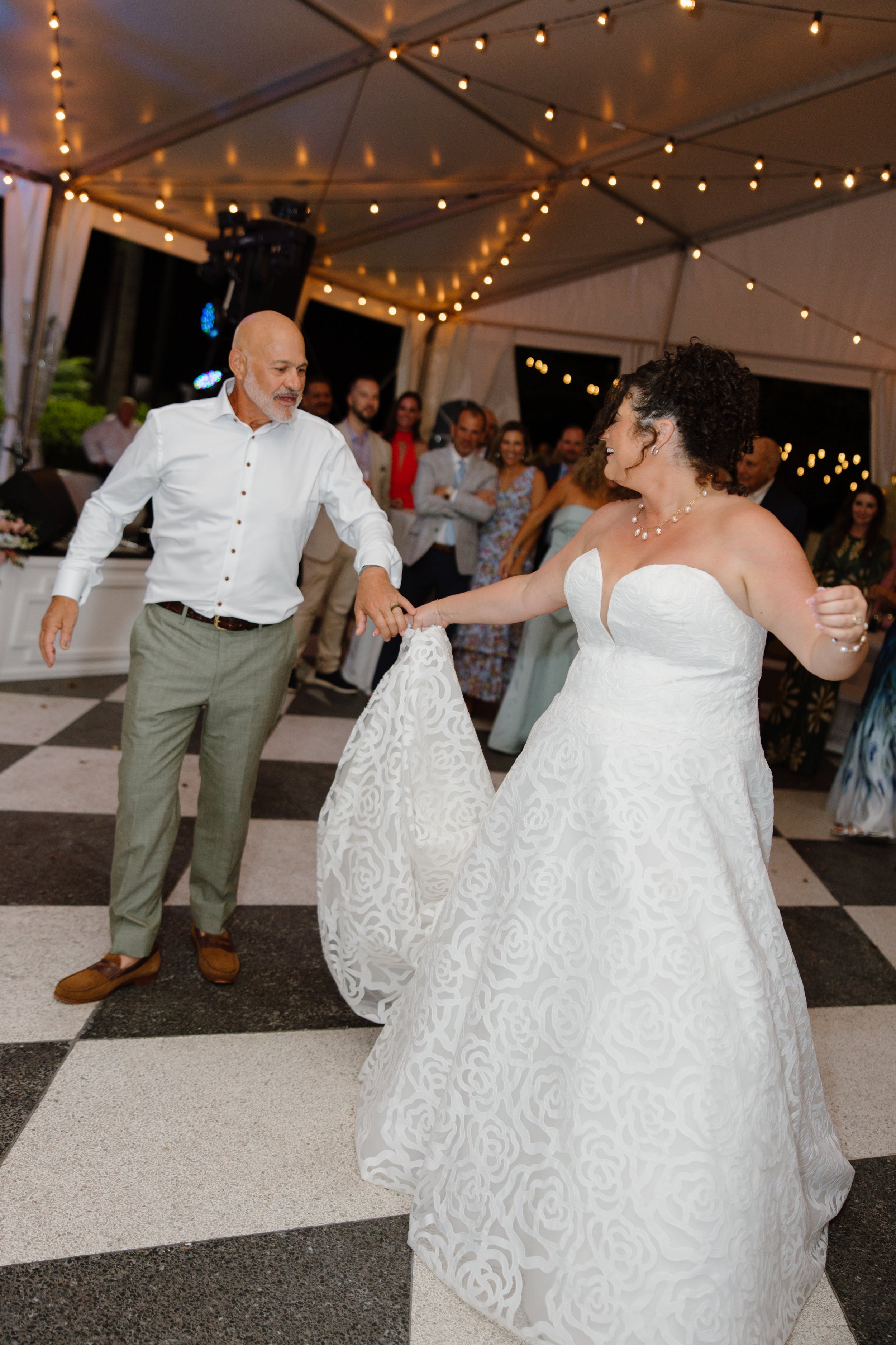 A wedding reception with a bride in a white wedding dress dancing with an older man in a white shirt and gray pants, under a tent with string lights, and surrounded by guests in formal attire.