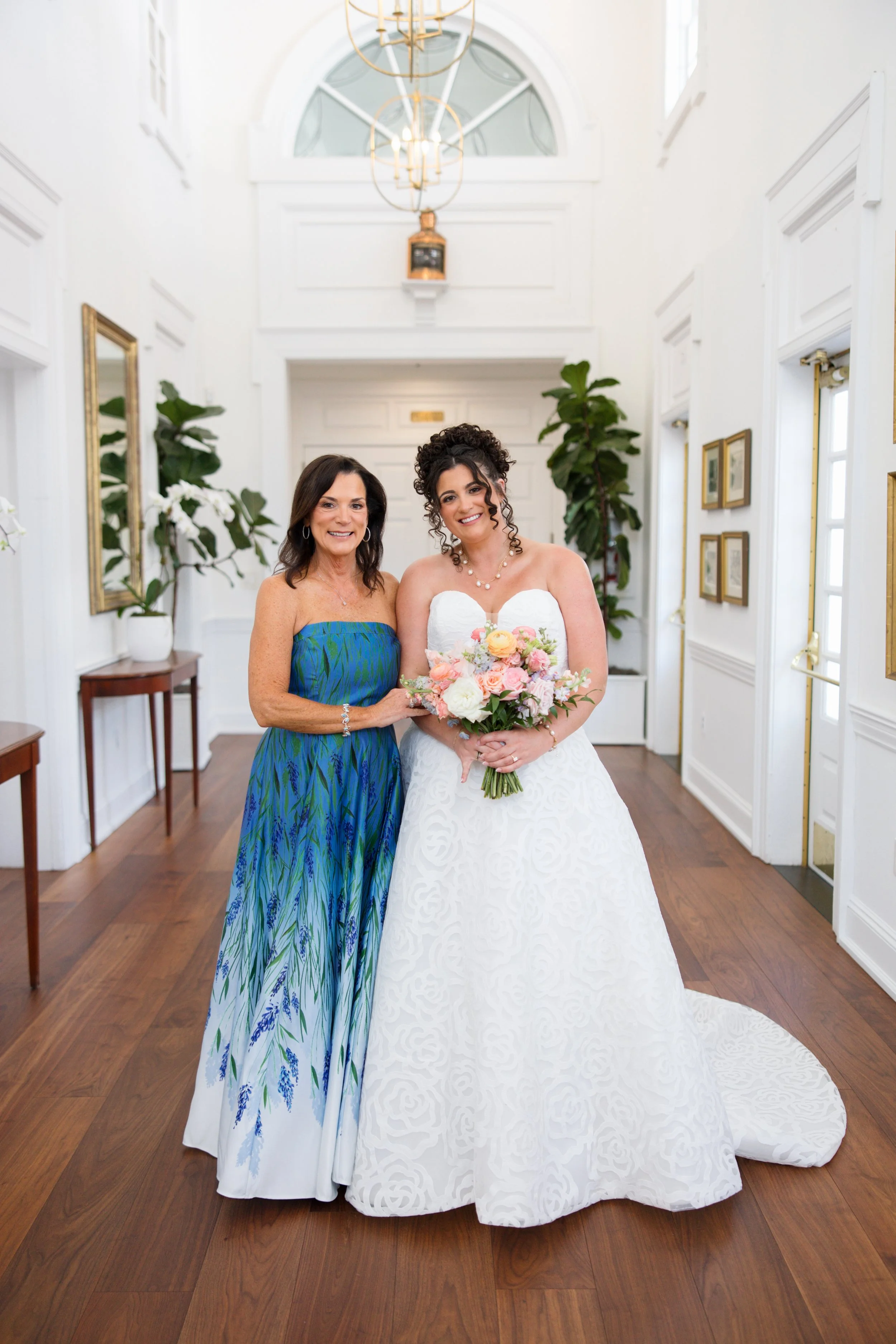 Two women standing in a bright, white, elegant room with wooden floors and large windows. One woman is dressed in a white wedding gown holding a bouquet, and the other is wearing a strapless blue and green patterned dress. Both are smiling at the cam