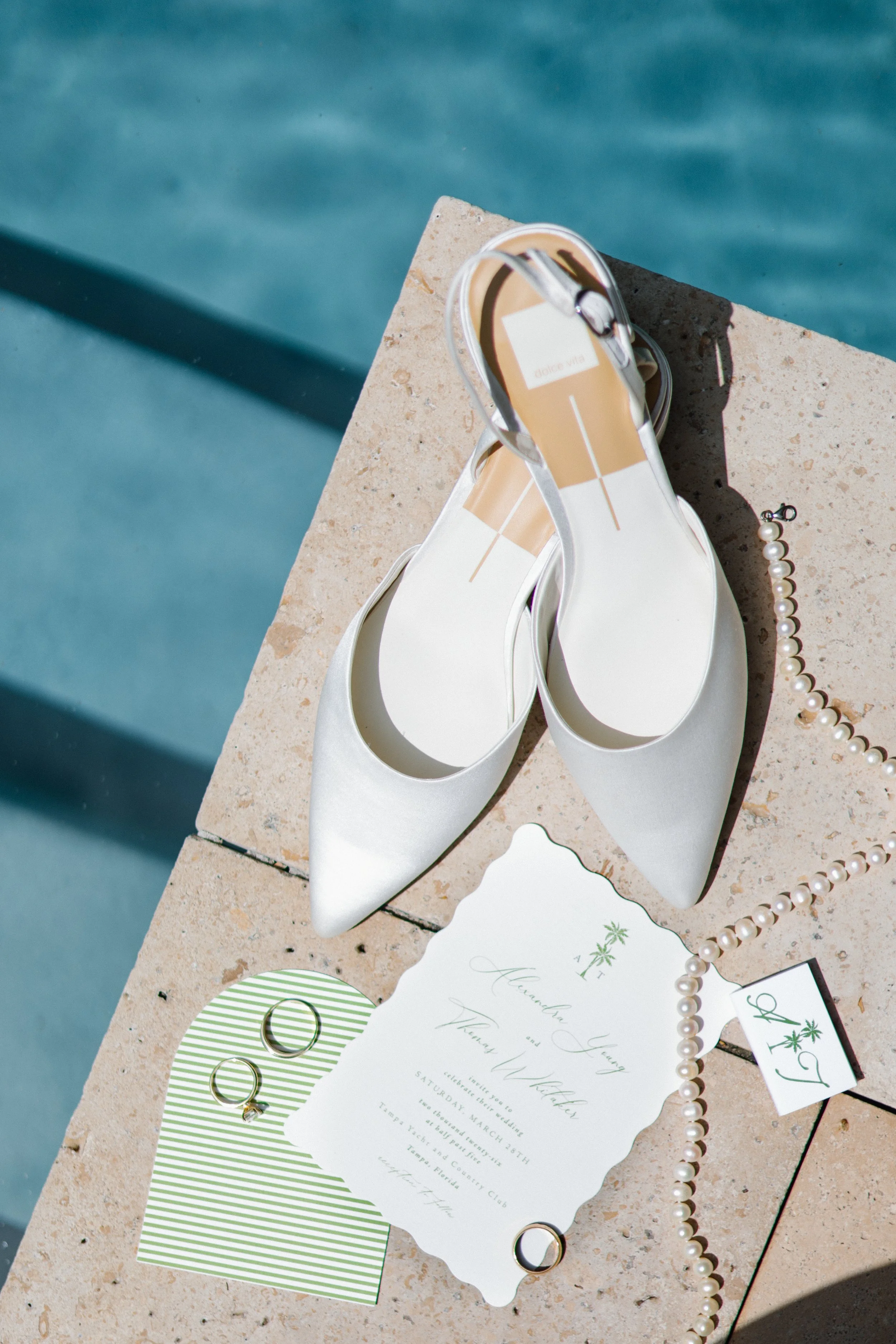 White high-heeled shoes with clear straps, pearl necklace, wedding invitation cards, and three gold rings on a textured beige surface next to a swimming pool.