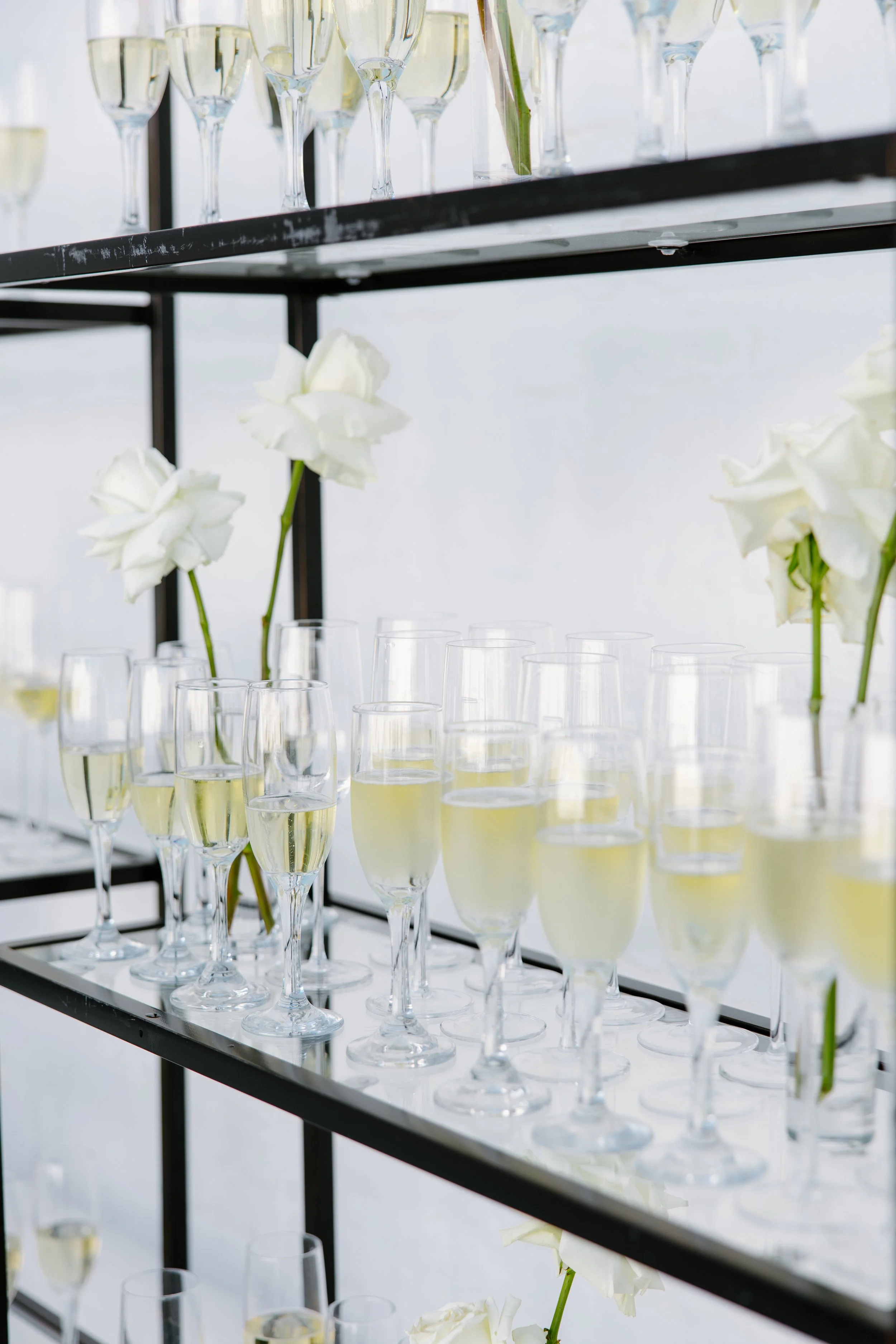 Multiple champagne flutes filled with champagne and white roses in vases on a black metal stand, with a white background.