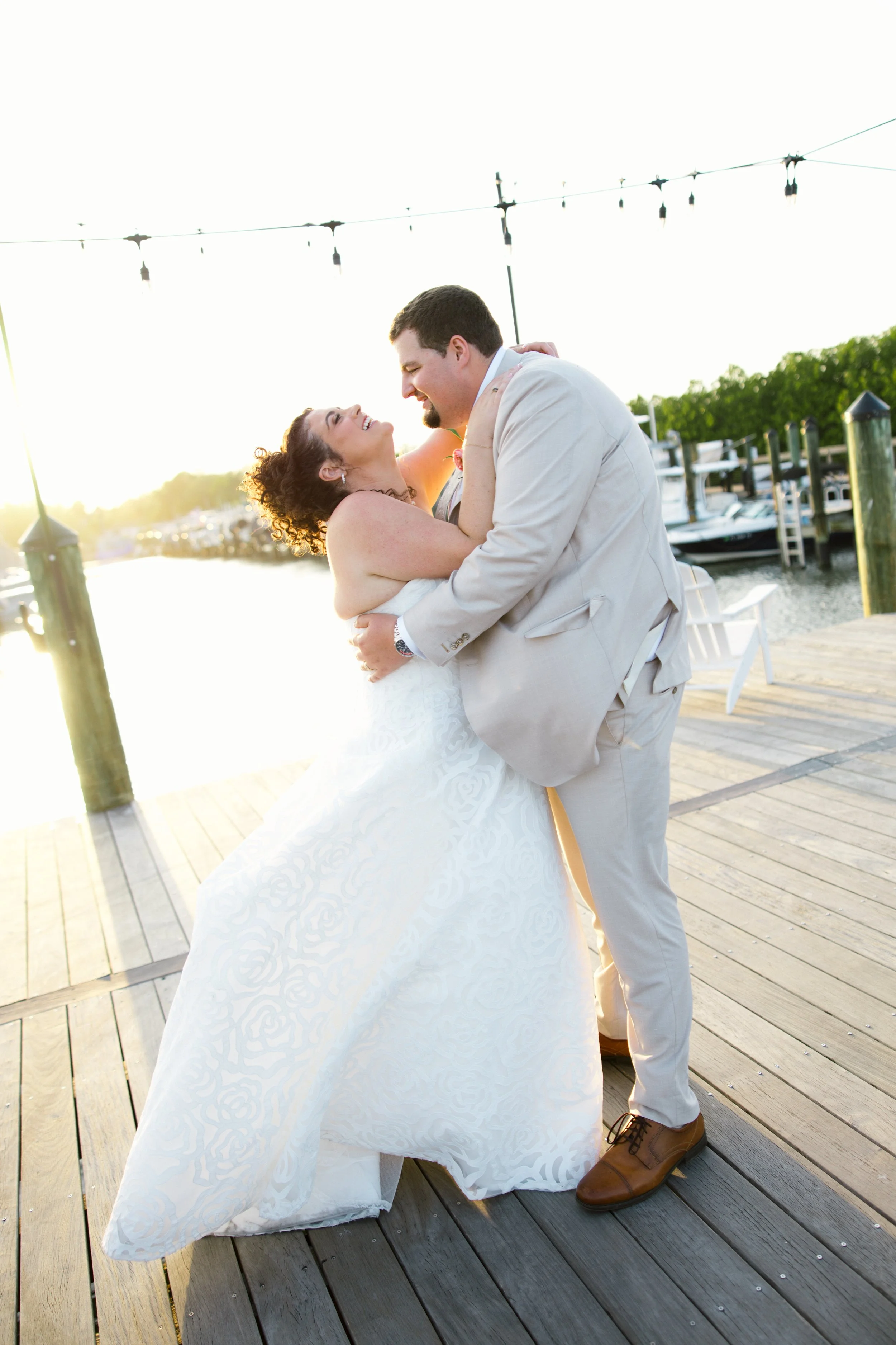 A bride and groom dancing on a wooden dock at sunset, with boats and a marina in the background.