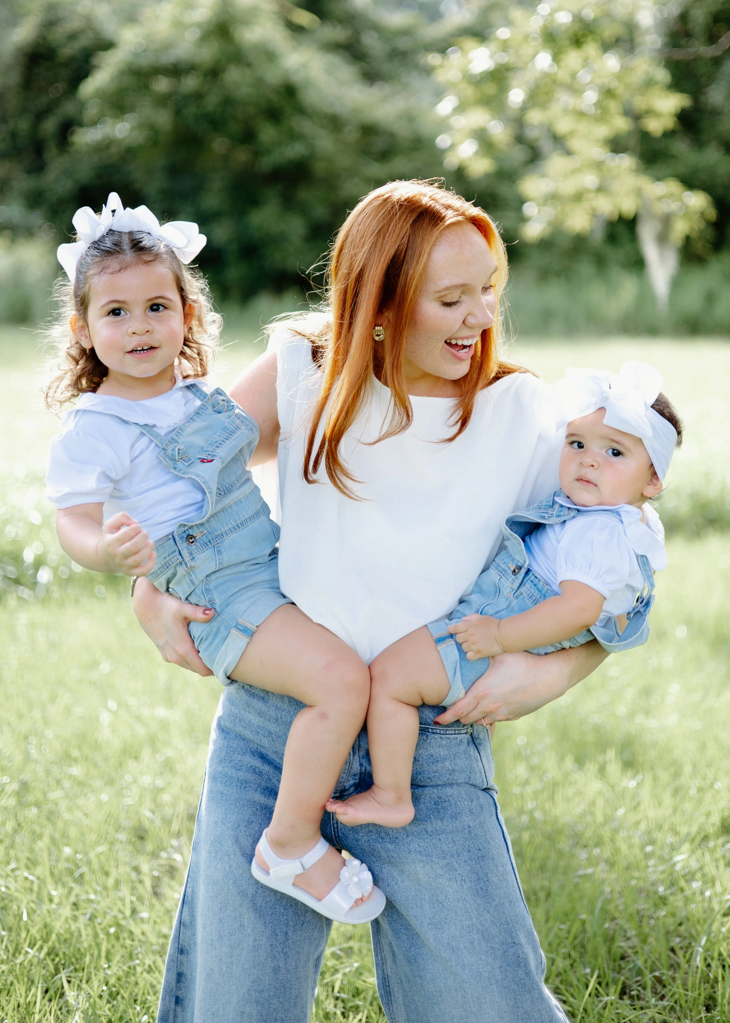A woman with red hair smiling and holding two young girls, all wearing white tops and denim overalls, outdoors in a grassy park with trees in the background.
