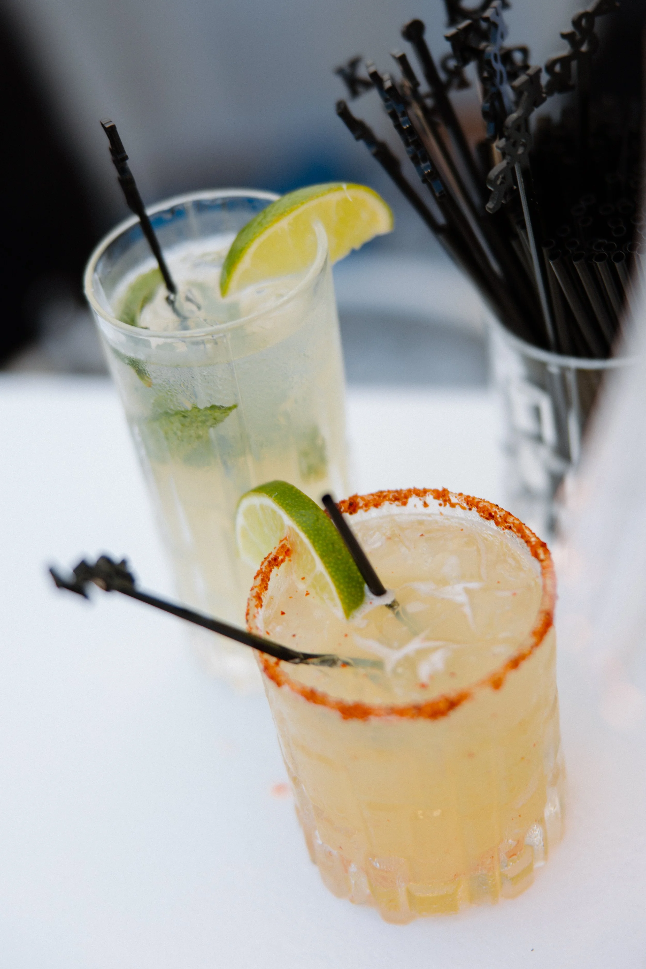 Close-up of a chili rimmed cocktail glass with a lemon wedge, filled with a light-colored drink, in front of a container holding black drinking straws.