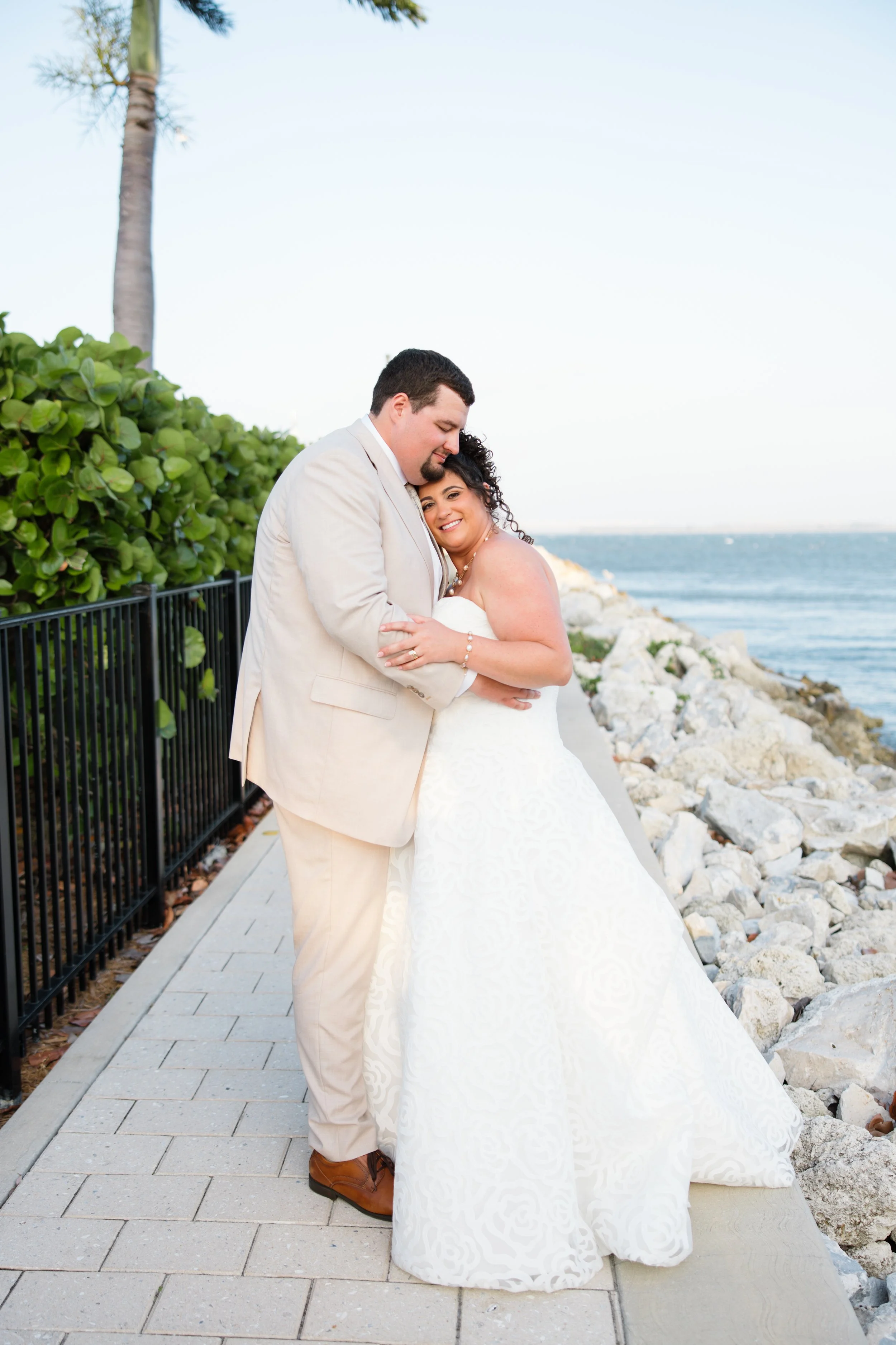 A newlywed couple standing close together on a seaside pathway, with the man holding the woman, who is smiling, as they pose for a wedding photo. The woman is in a strapless white wedding gown, and the man in a light beige suit. The background featur