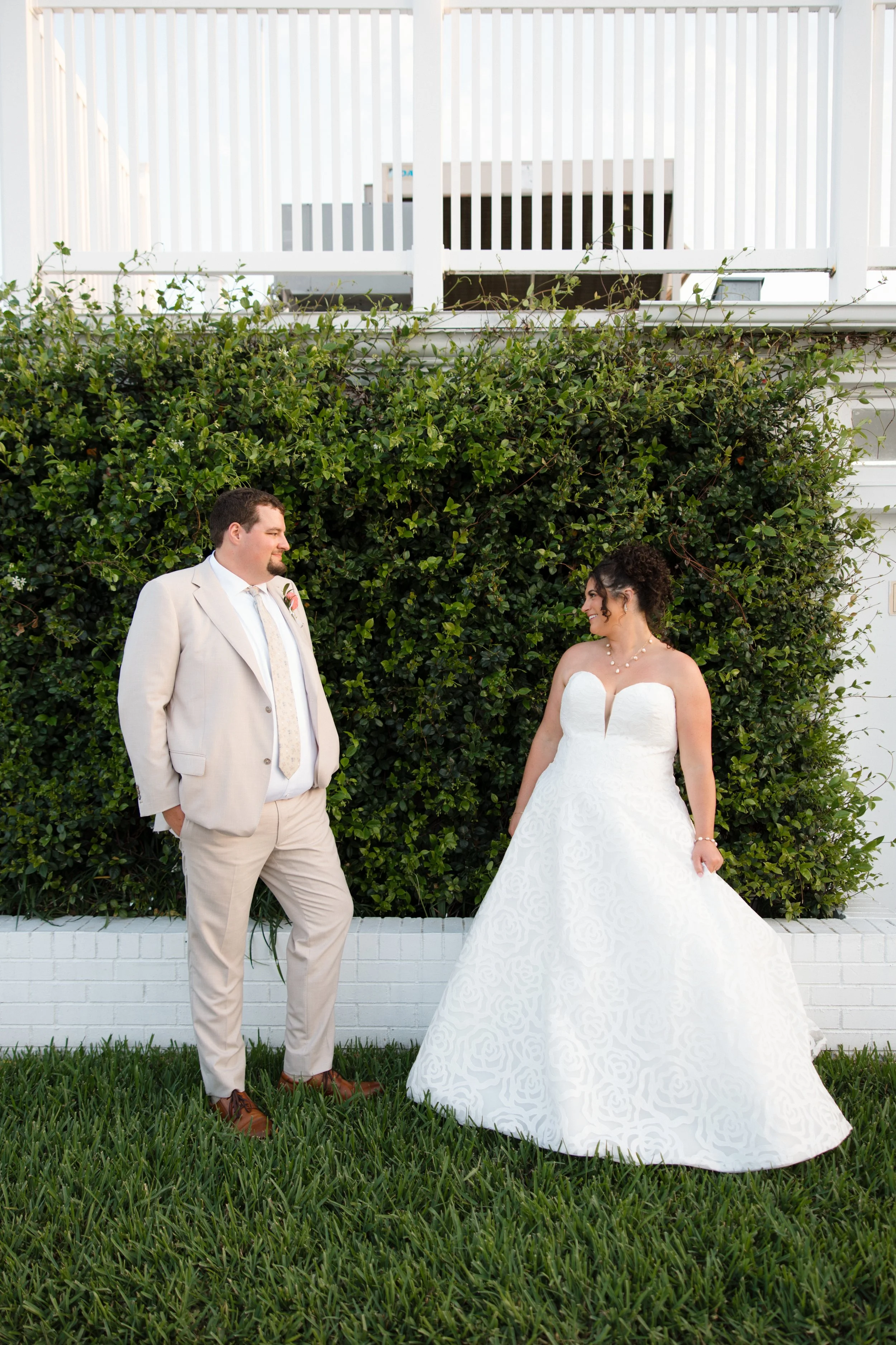 A bride and groom standing outdoors in front of a green hedge with white fencing above. The groom, wearing a light beige suit, is smiling at the bride, who is wearing a strapless white wedding gown. They are looking at each other, with the bride smiling broadly.