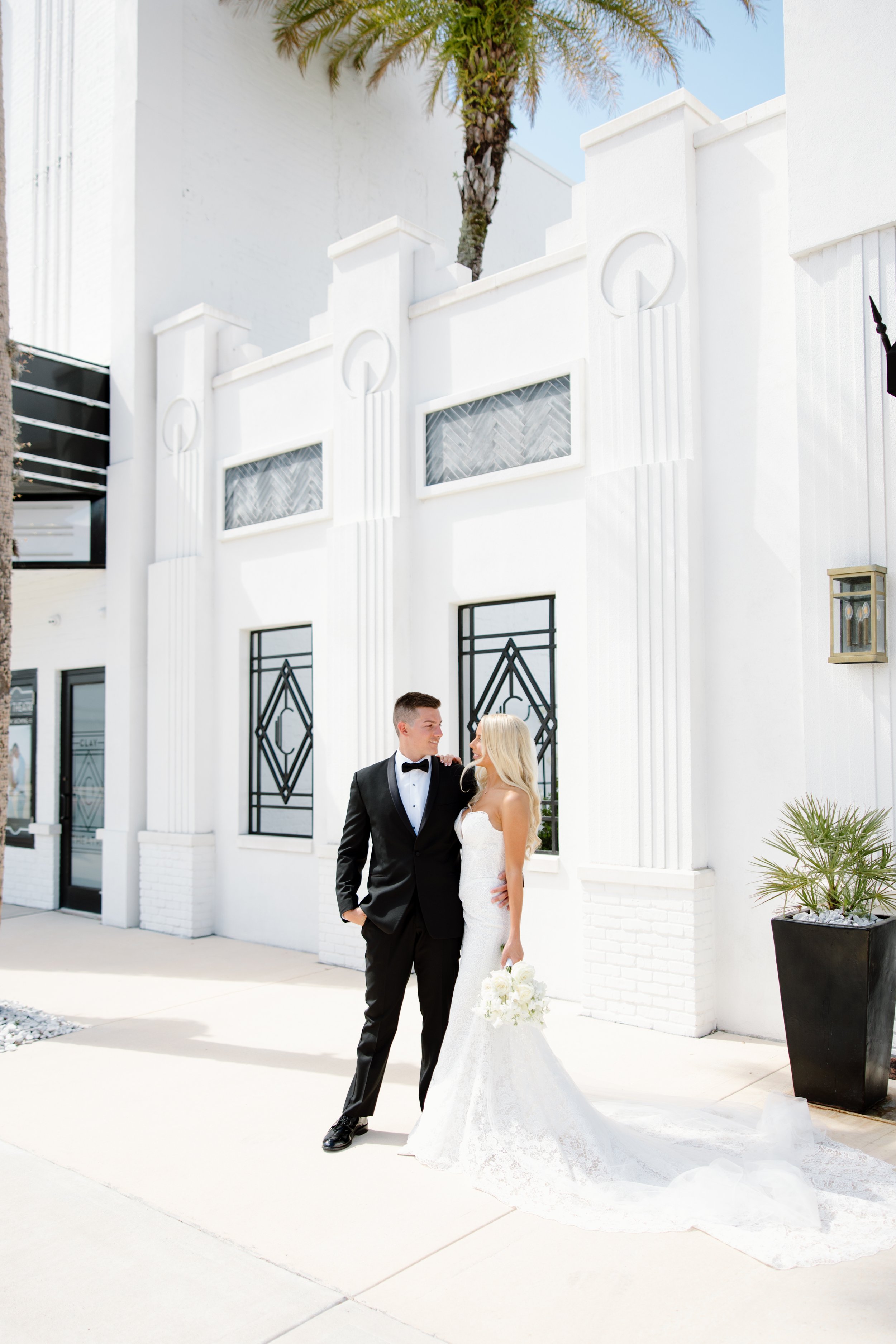 A newlywed couple stands outside a modern white building with black accents and palm trees in the background, sharing a tender moment.