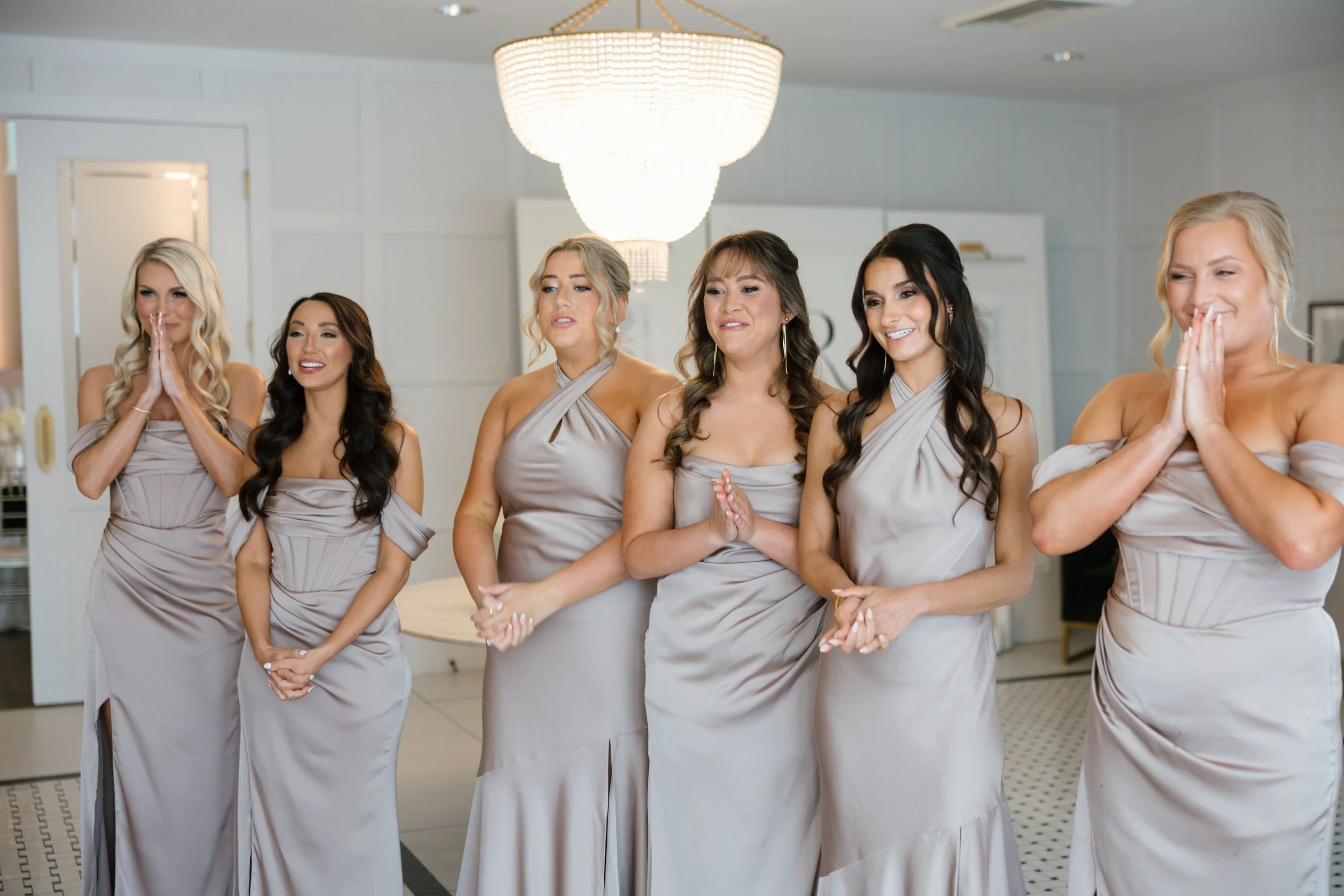 Six women in matching light purple dresses standing in a line indoors, some with hands clasped and some with hands near their faces, smiling and showing various expressions.