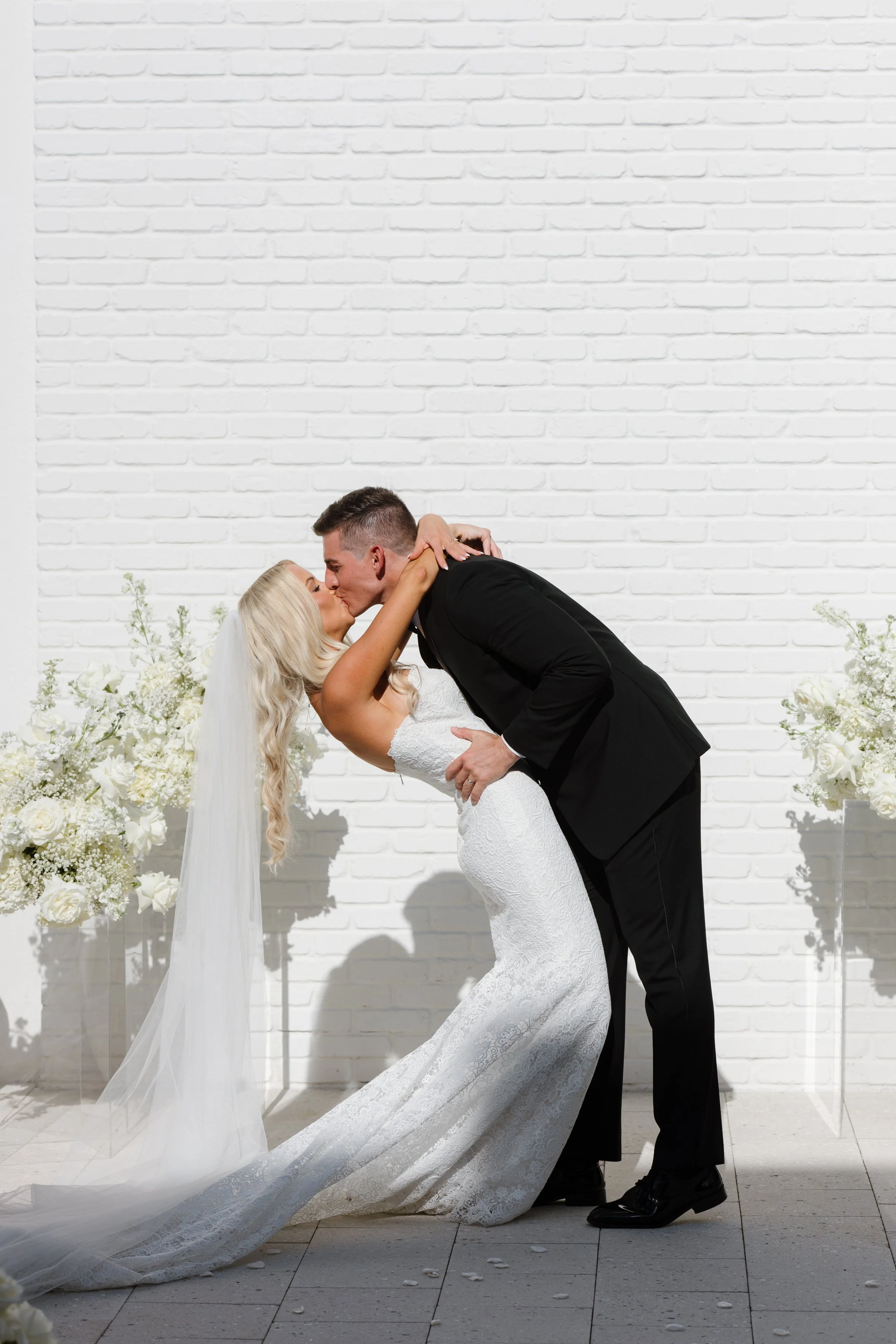 A bride and groom share a kiss during their wedding ceremony, with the bride leaning back in the groom’s arms, against a white brick wall with floral arrangements.