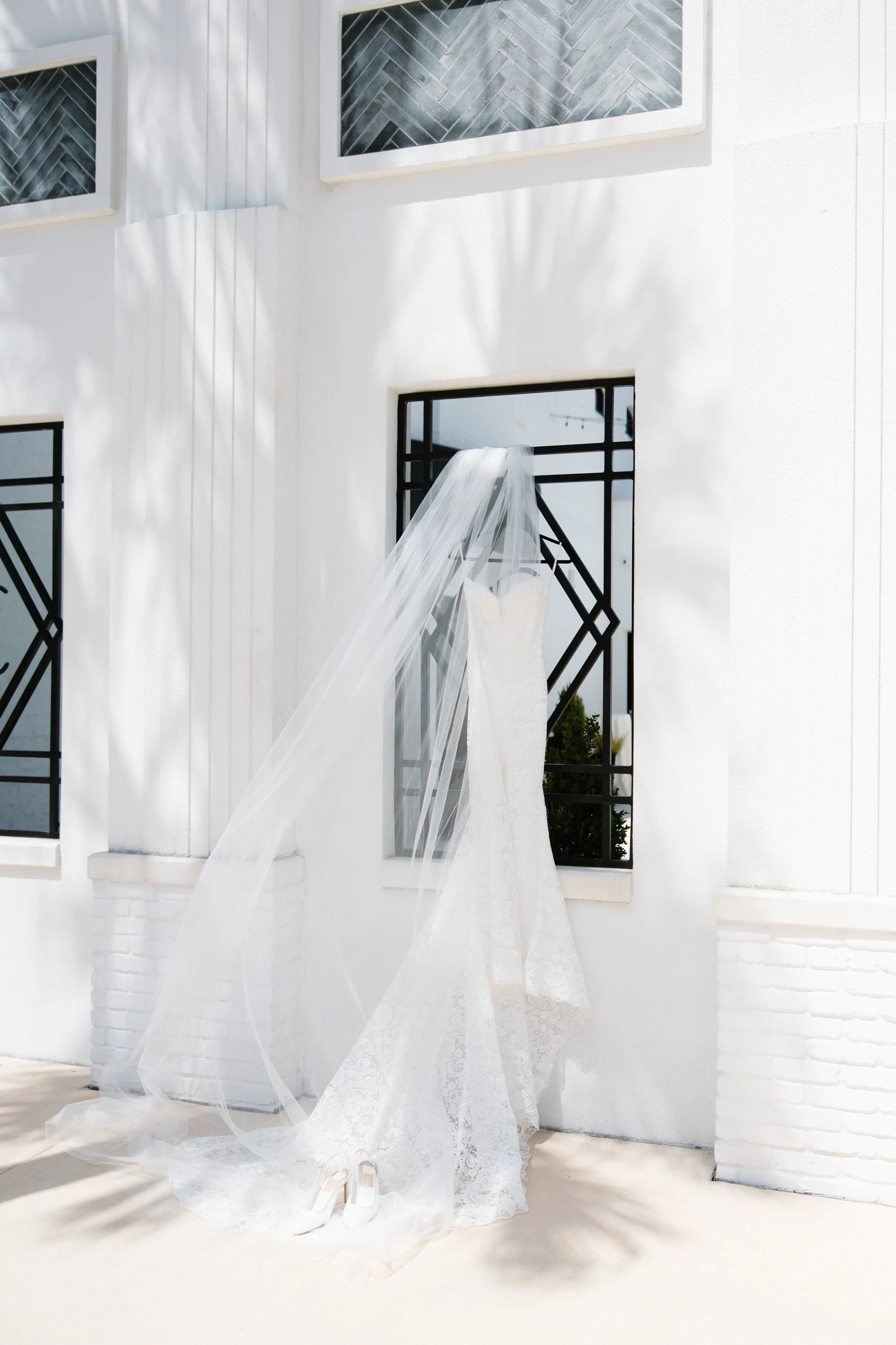 White wedding dress and veil with white high-heeled shoes in front of a white wall and black window frames with geometric designs.