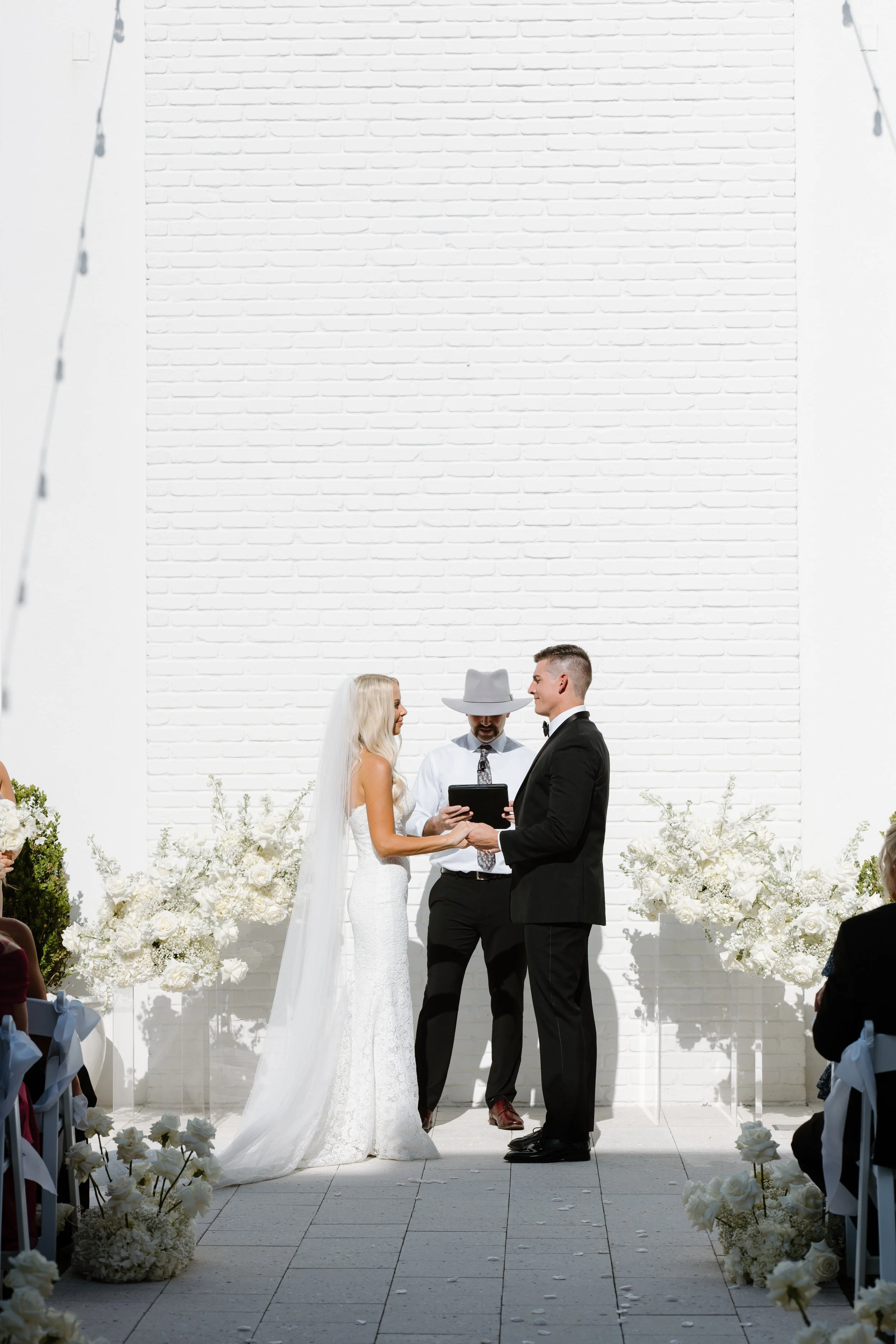 A bride and groom standing together during a wedding ceremony, facing each other and holding hands, with an officiant reading from a book, all dressed formally, outdoors with white floral arrangements and a white brick wall backdrop.