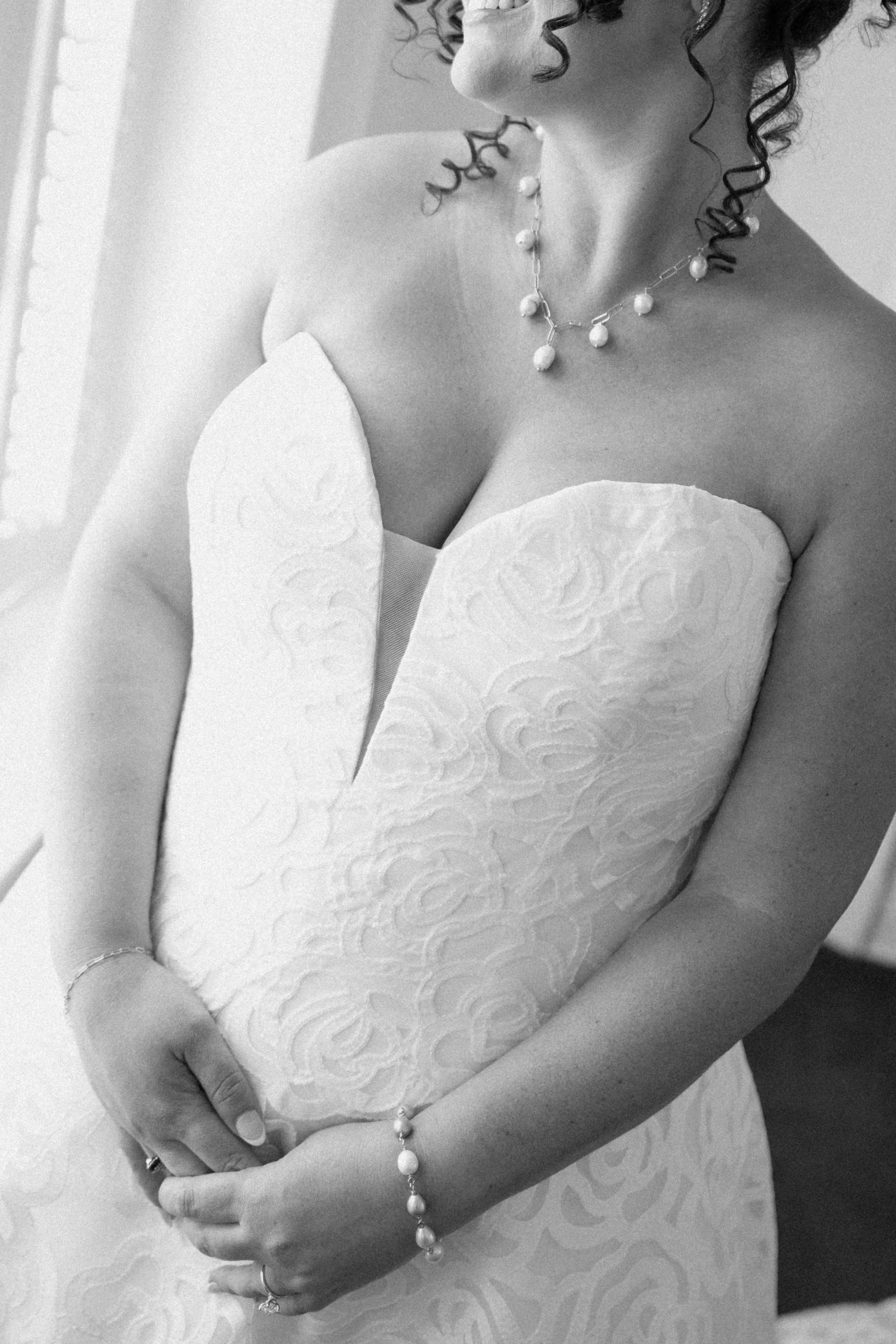 A woman wearing a white strapless dress with an intricate pattern, a pearl necklace, a bracelet, and rings, holding her hands over her abdomen, with curly hair and a smile partially visible.