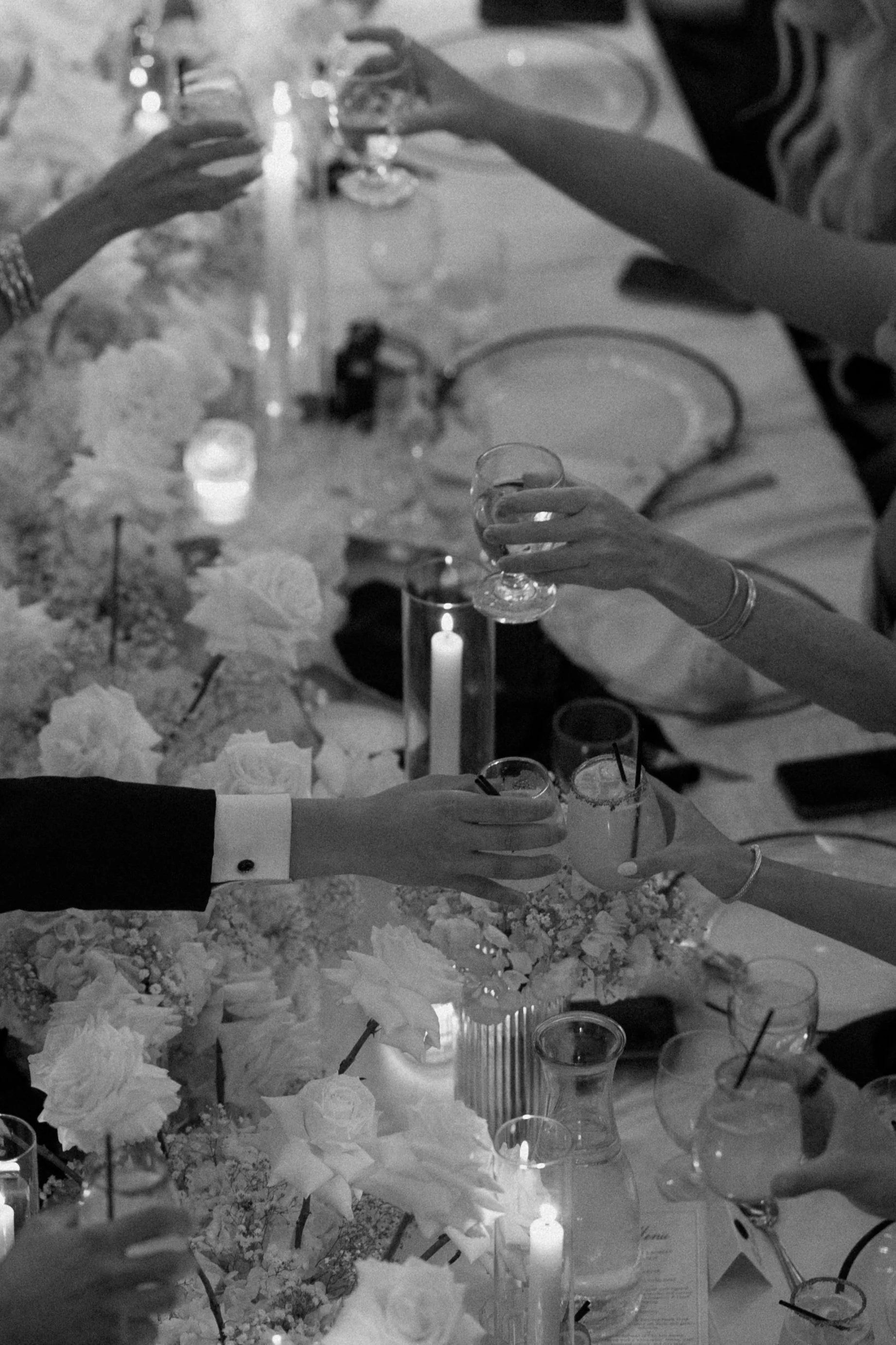 People raising glasses for a toast at a formal event, with floral centerpieces and lit candles on the table.