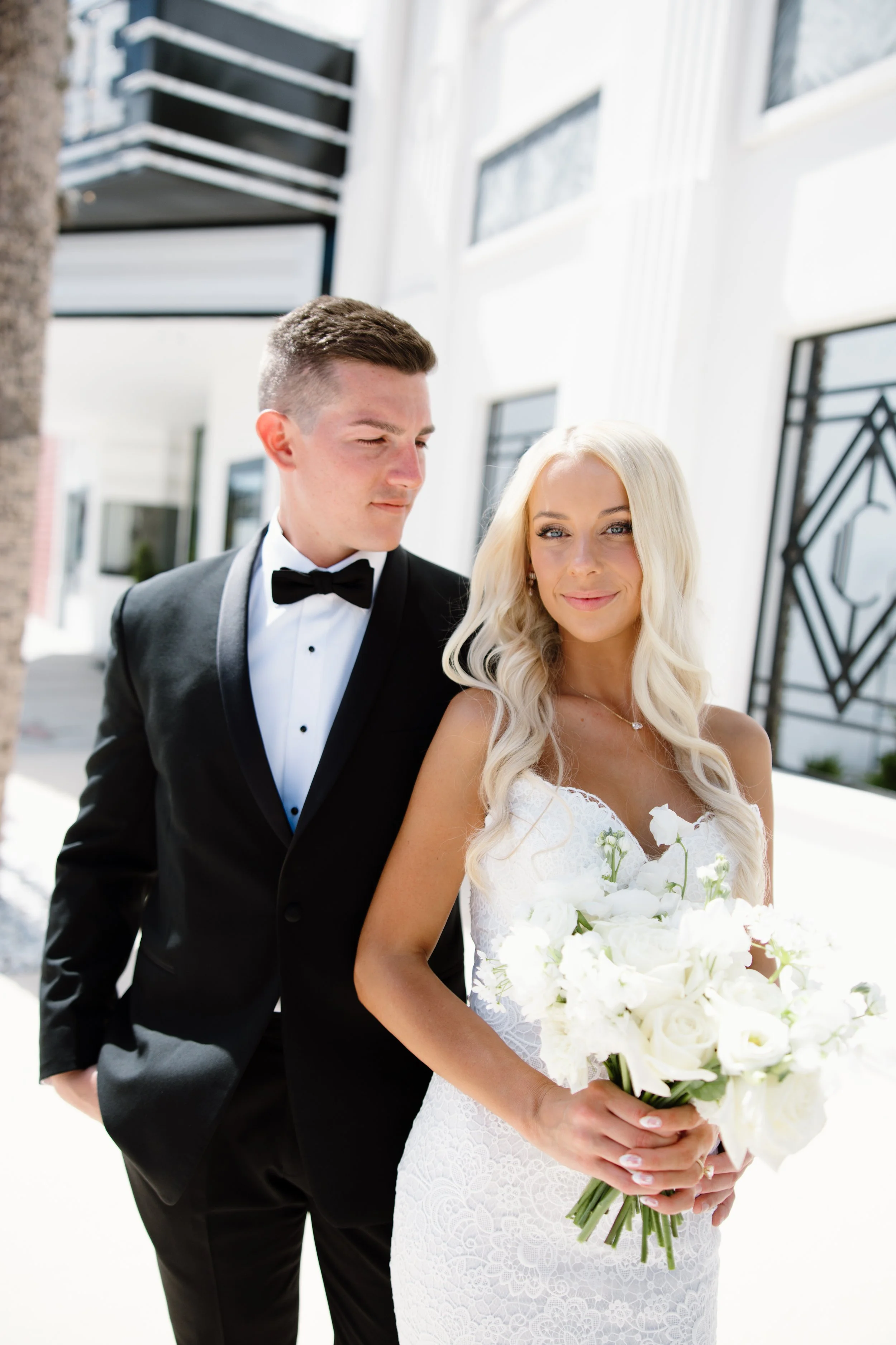 A bride with long blonde hair holding a bouquet of white flowers, wearing a white lace wedding dress, standing next to a groom with short dark hair in a black tuxedo, outdoors on a sunny day.