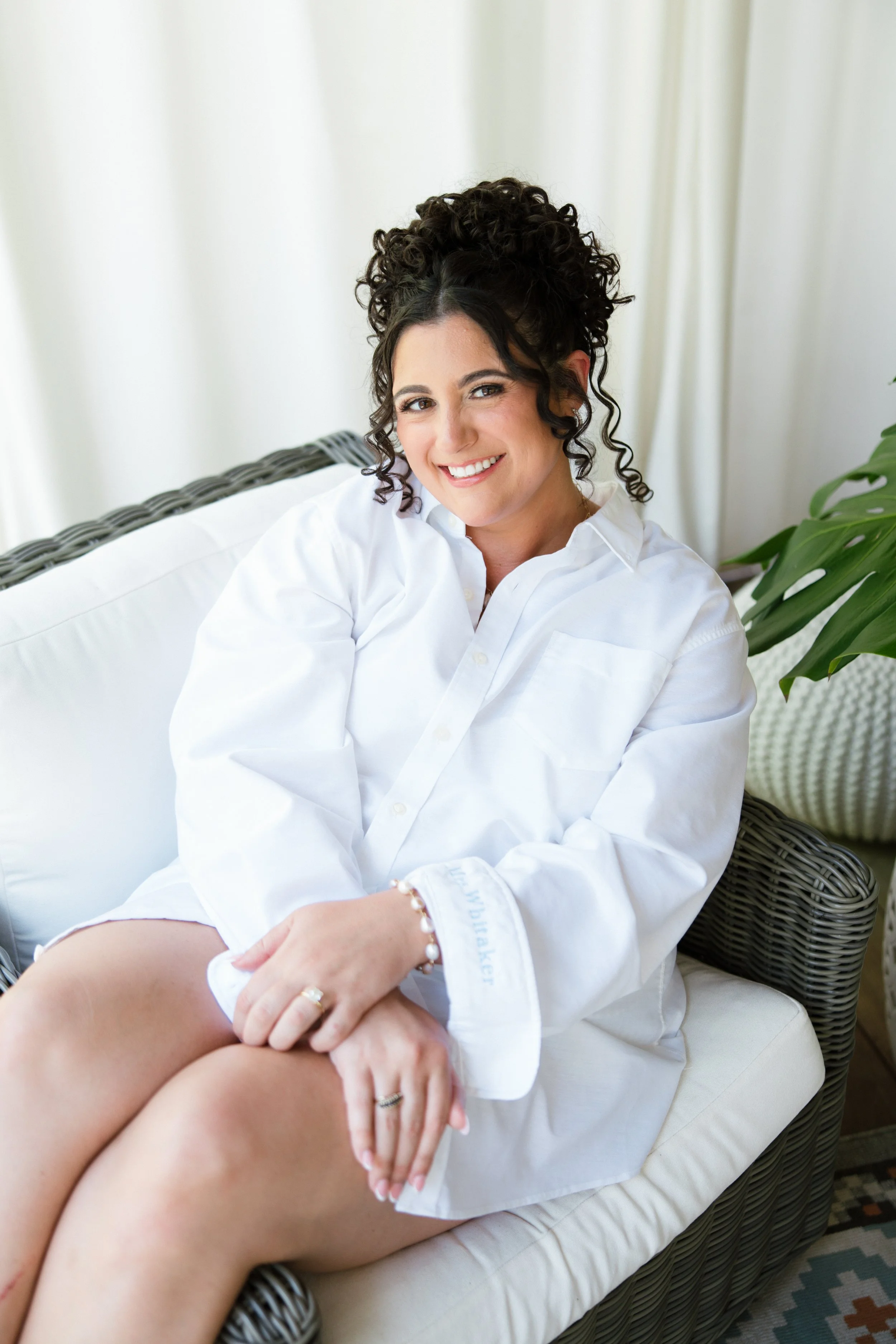 A woman with curly dark hair and a white shirt sitting on a wicker furniture, smiling at the camera, next to a green plant and white curtains in the background.