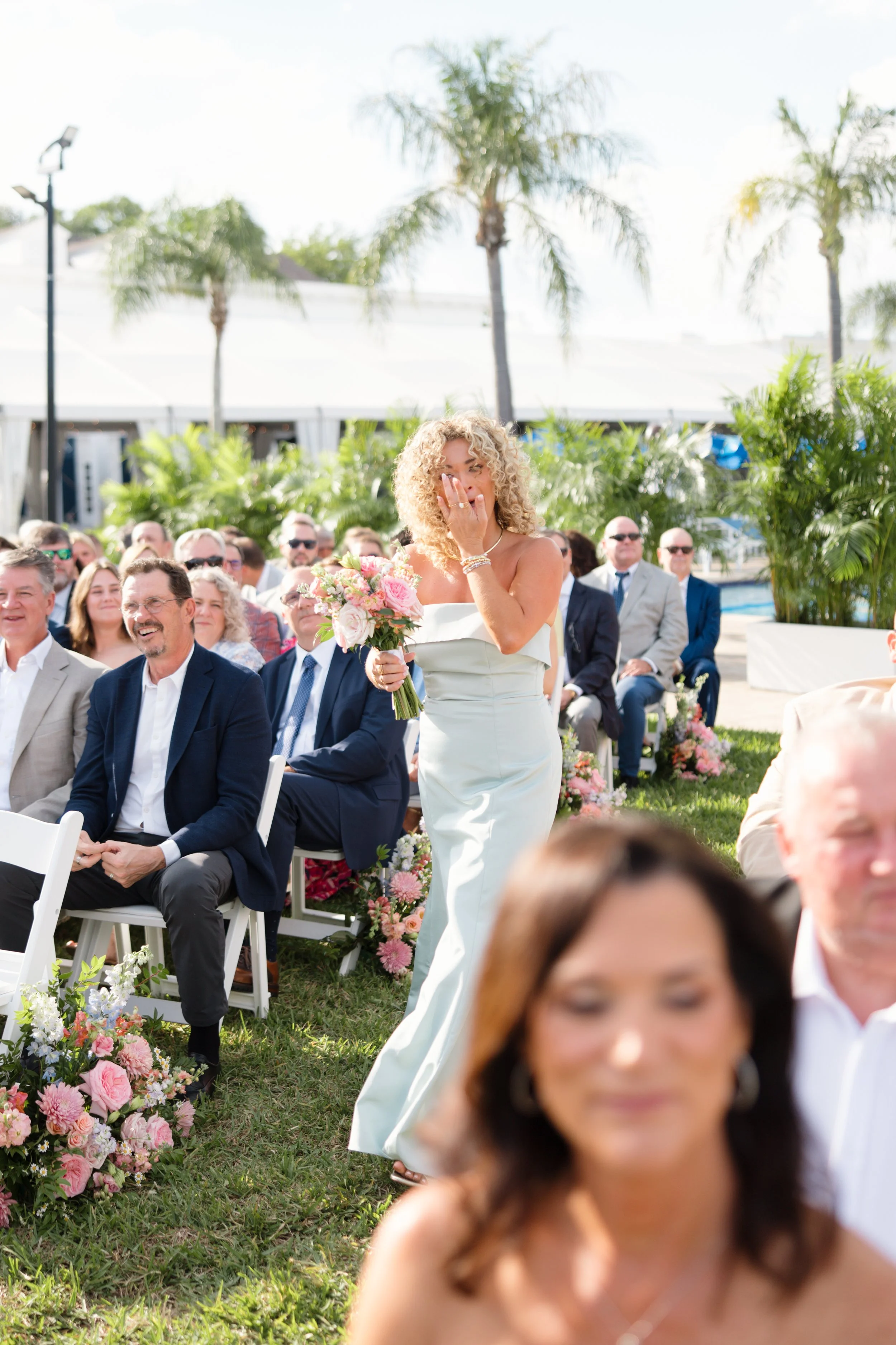 A woman in a white strapless dress holding a bouquet of pink and white flowers covers her face with her hand during an outdoor wedding ceremony, with seated guests and palm trees in the background.