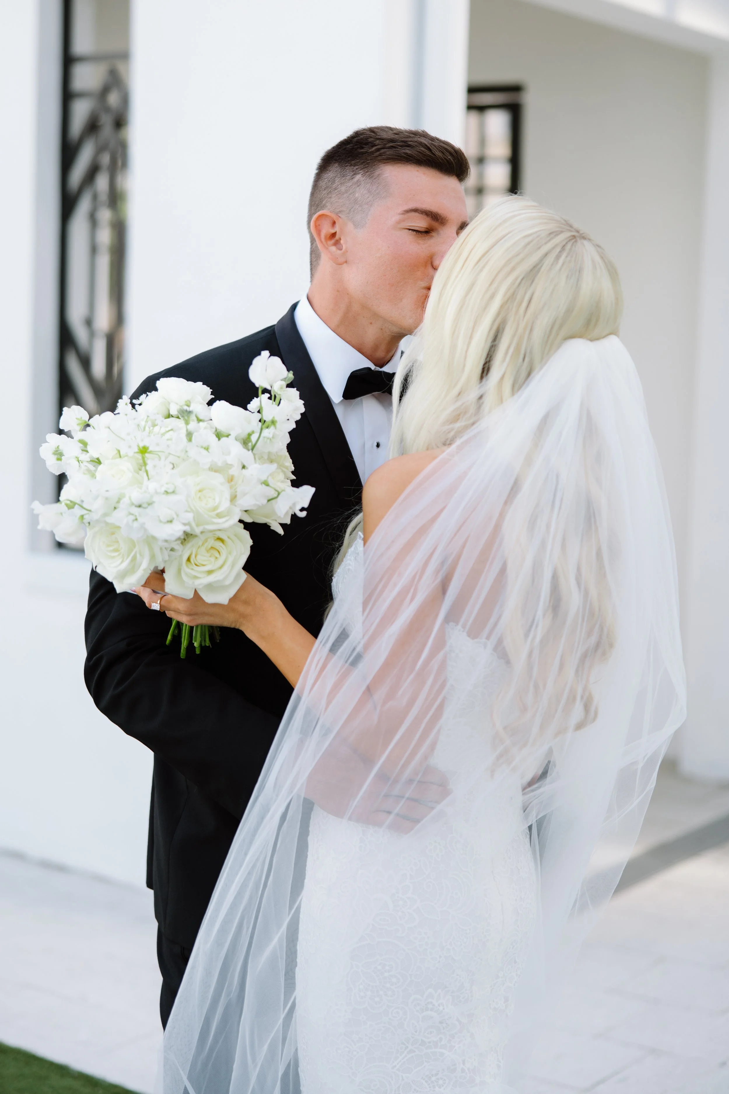 A groom in a black tuxedo and bow tie kissing a bride in a white wedding dress with a veil, holding a bouquet of white flowers, outside a modern building.