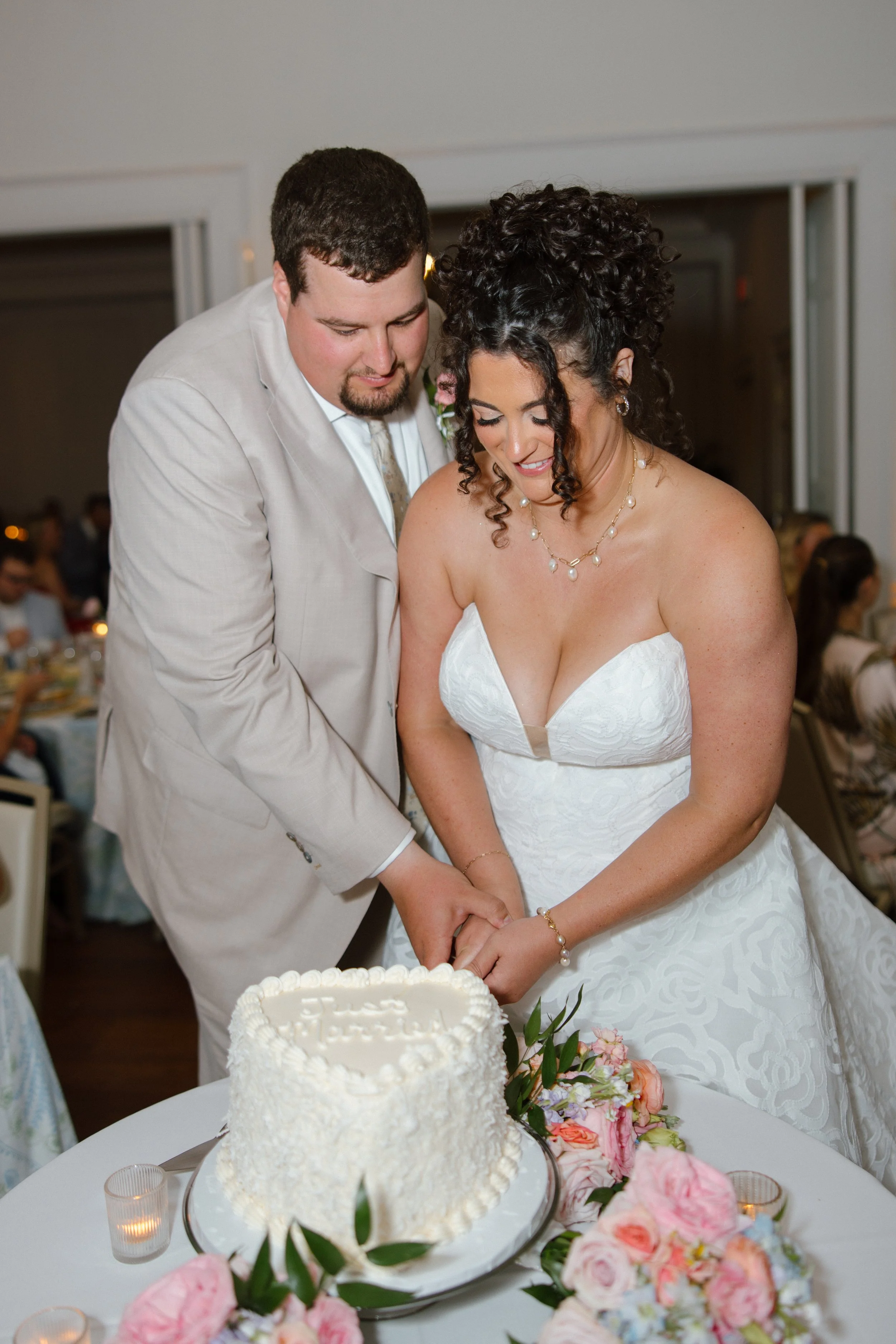 A bride and groom cutting a wedding cake at their reception, surrounded by flowers and guests.