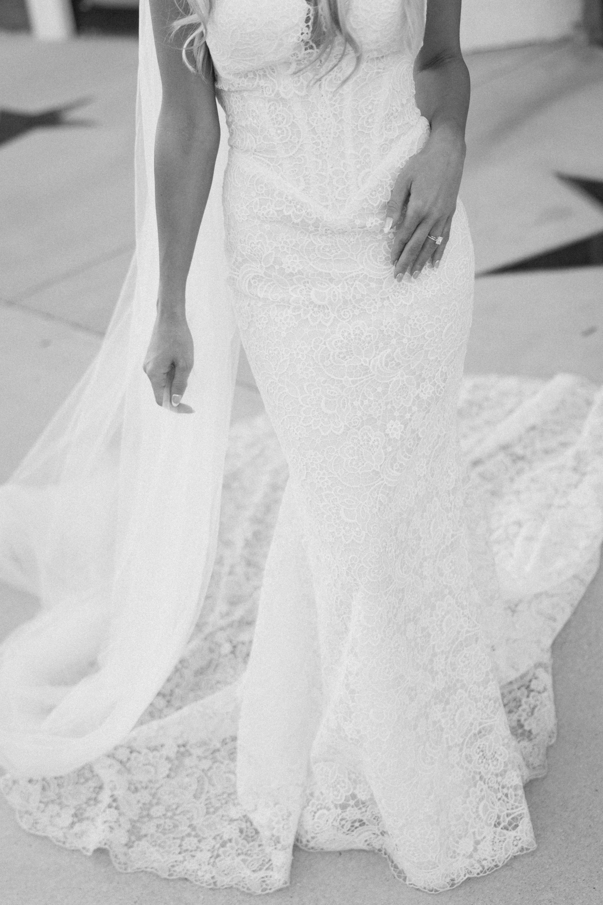 Black and white photo of a woman wearing an elegant lace wedding dress, with her left hand resting on her hip, showing her wedding ring.