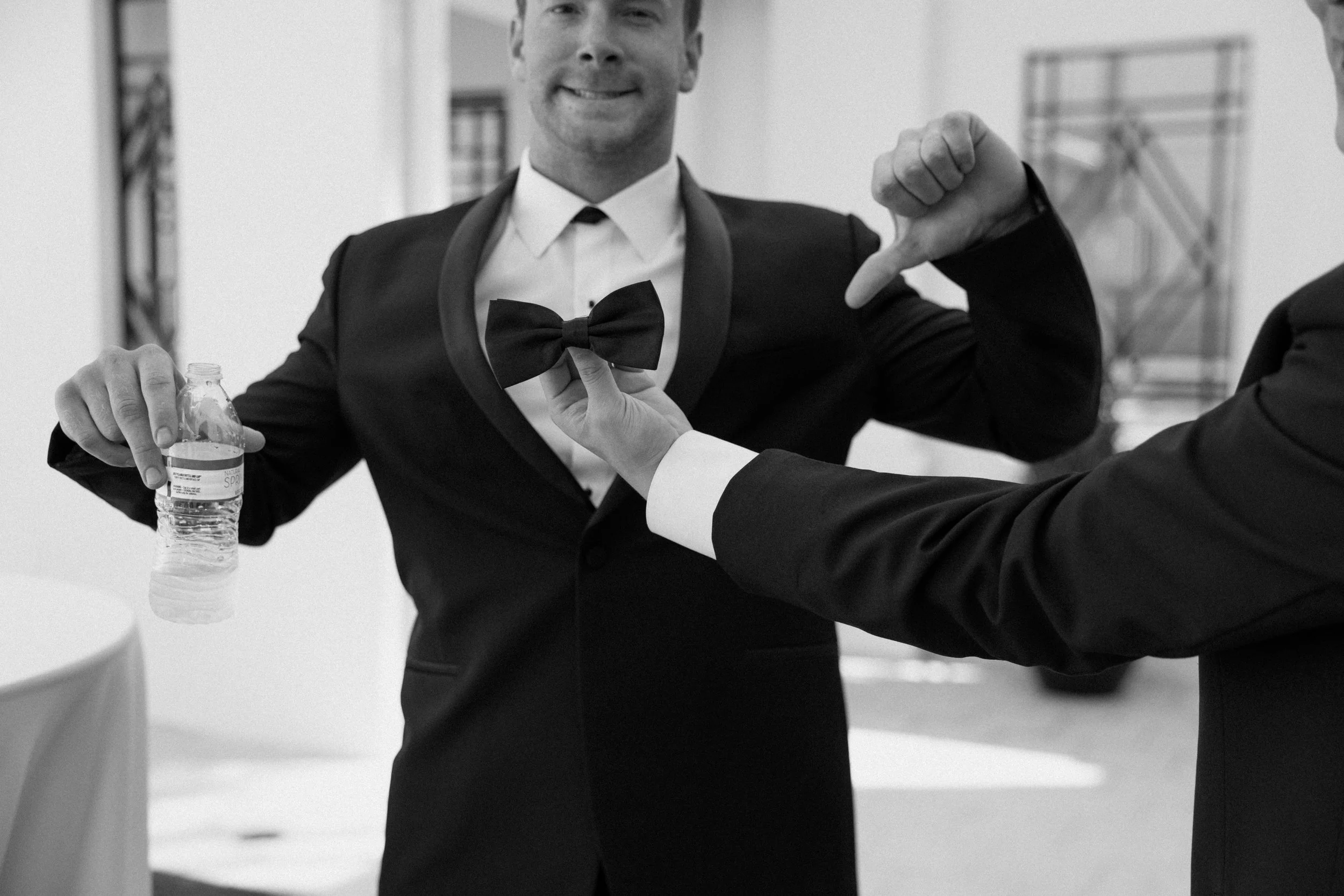 A man in a tuxedo smiling and pointing at an open shirt button while another person holds a water bottle, in a black-and-white photo.