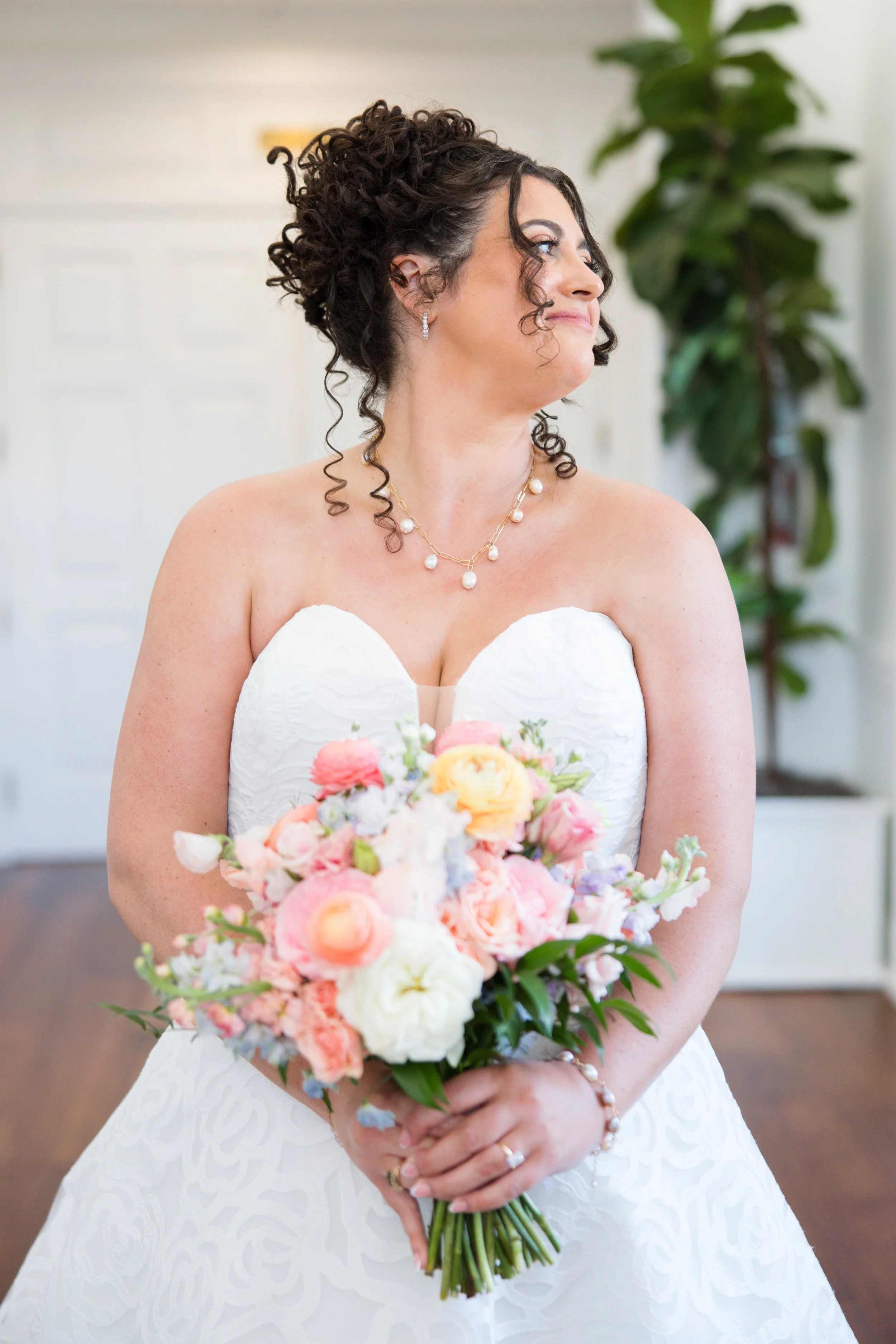 A woman in a white wedding dress holding a bouquet of pink, white, and peach flowers in front of her with both hands, showing jewelry including earrings, a necklace, a ring, and a bracelet, standing indoors with a green potted plant and a white wall 