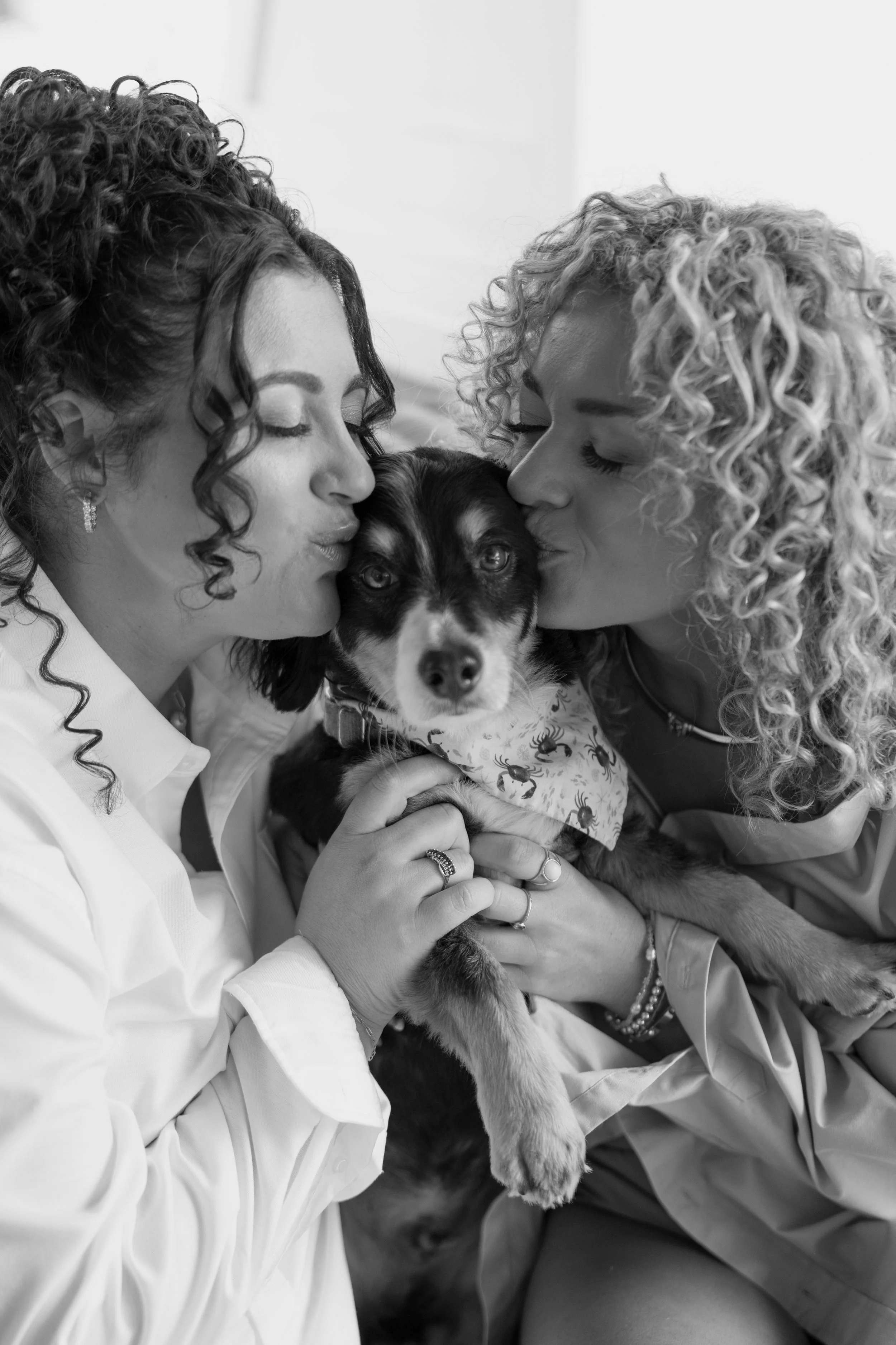 Two women with curly hair and earrings kissing a dog on the nose. The dog is wearing a patterned bandana.