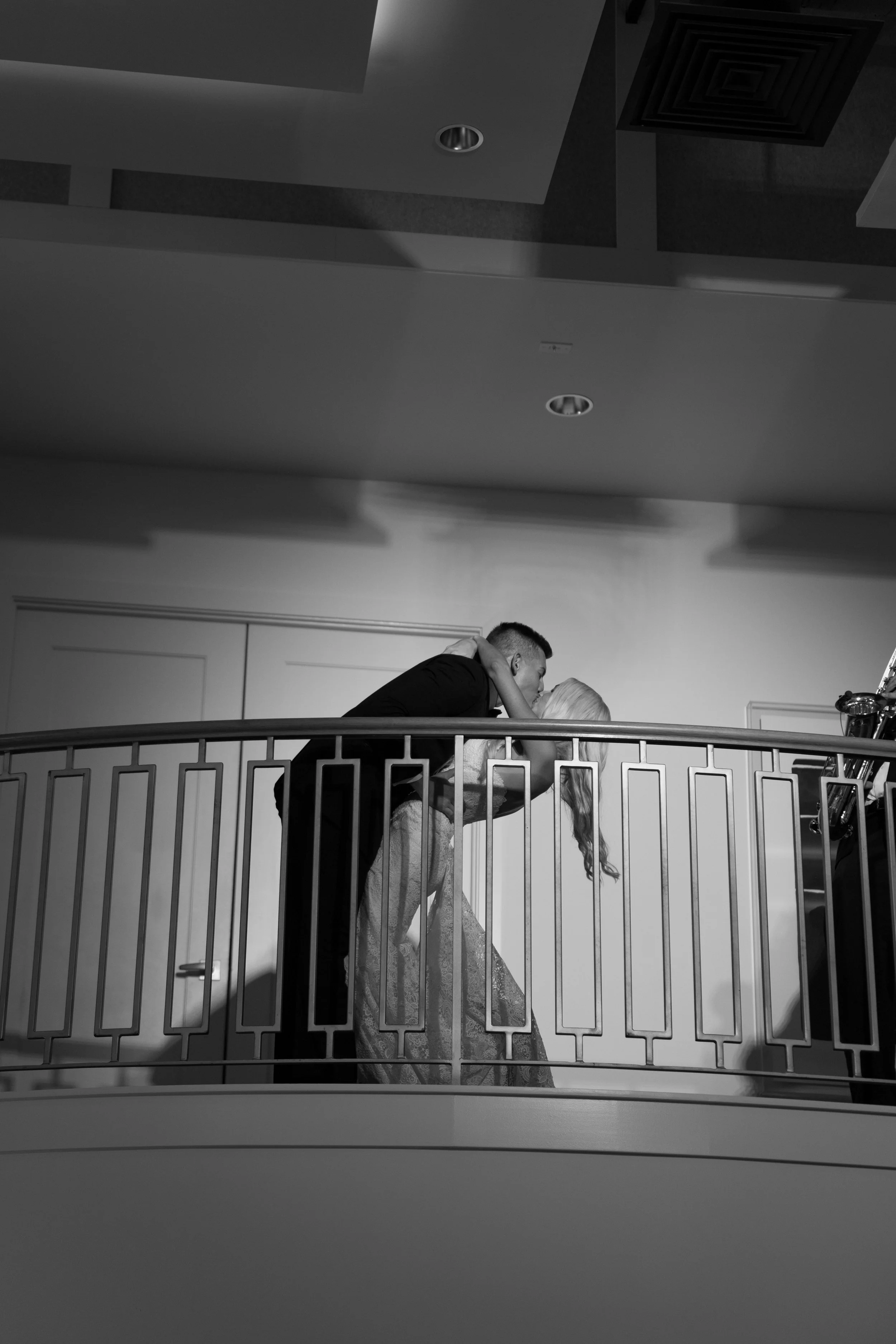 Black and white photo of a couple kissing on an indoor balcony, with the man leaning over the railing and the woman holding his shoulders, viewed from below.