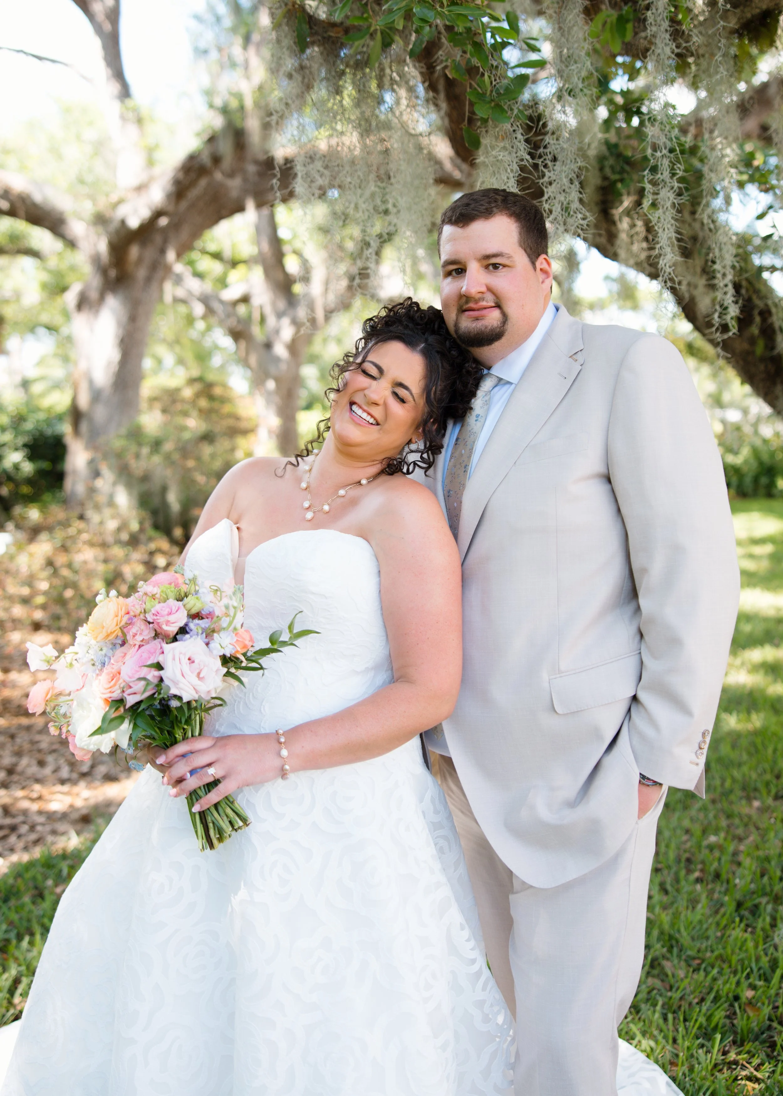 A bride and groom standing outdoors under a tree, smiling and embracing. The bride is holding a colorful bouquet and wearing a white wedding dress with a pearl necklace. The groom is dressed in a light gray suit and light blue shirt.