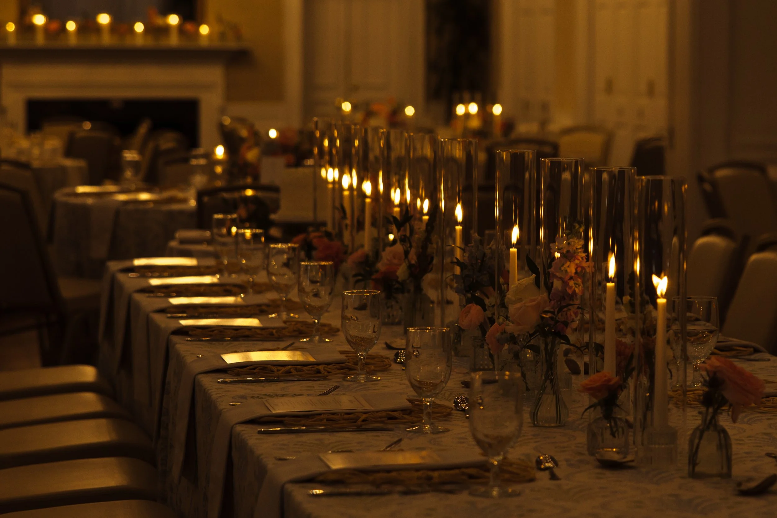 A long banquet table decorated with flowers and candles in glass holders, set for a formal dinner in a dimly lit room.