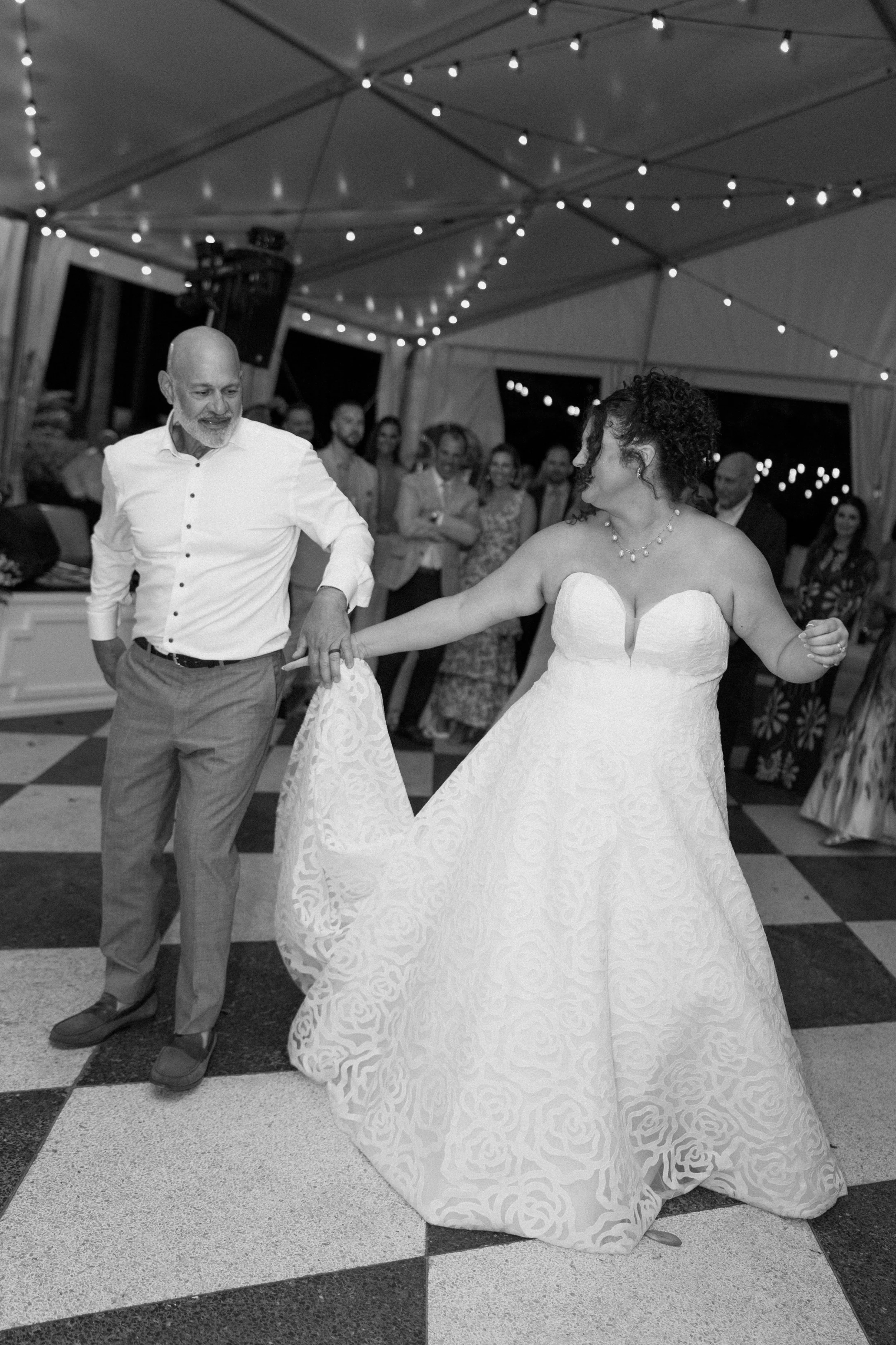 A bride and an older man are dancing at a wedding reception under string lights inside a tent. Guests are watching in the background.
