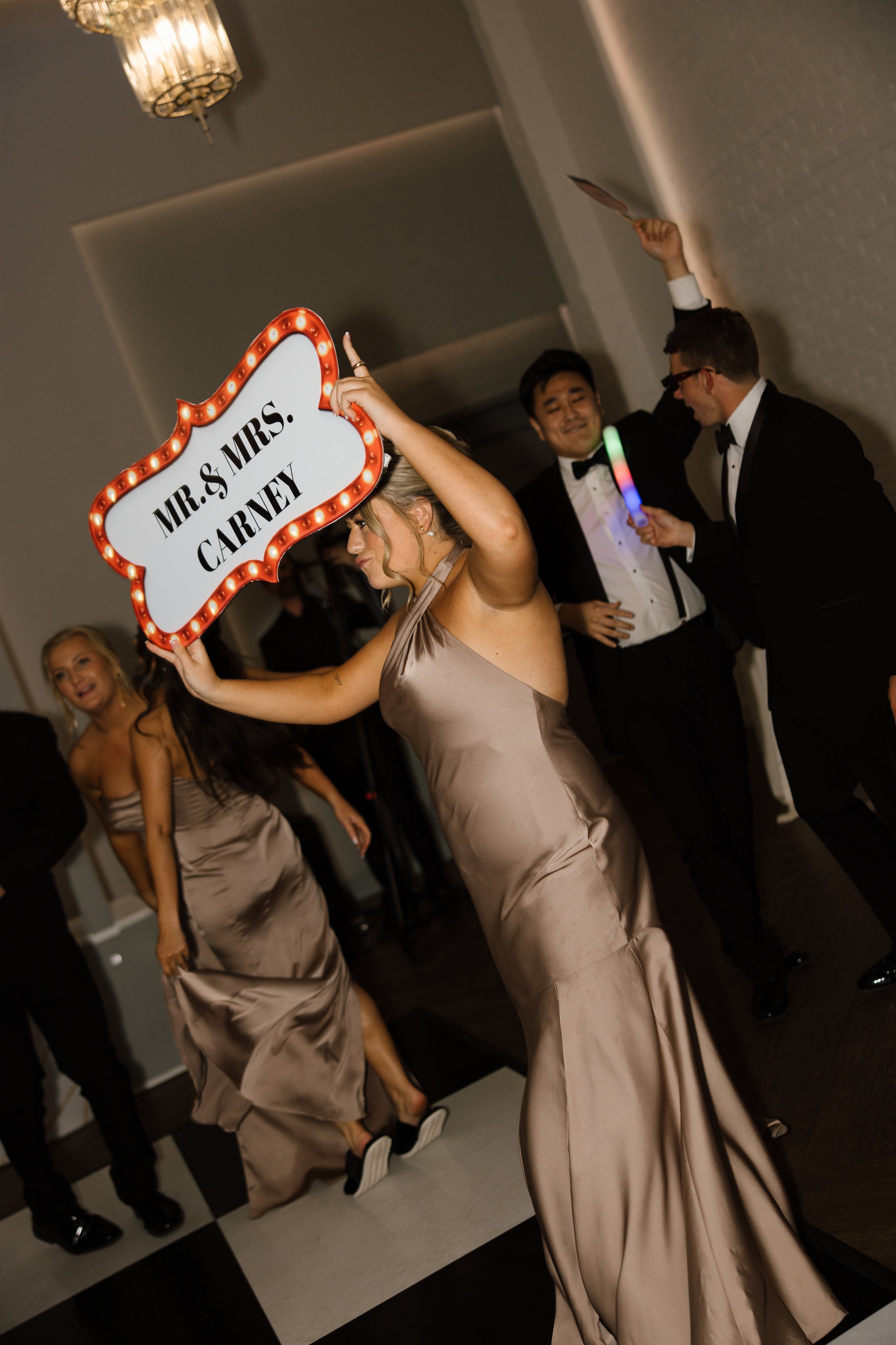 A woman in a beige satin dress at a wedding reception holding a sign that says 'Mr. & Mrs. Carney' with light bulbs around the border. She is smiling and pointing upwards. Other people in formal attire, including men in tuxedos, are dancing and celeb