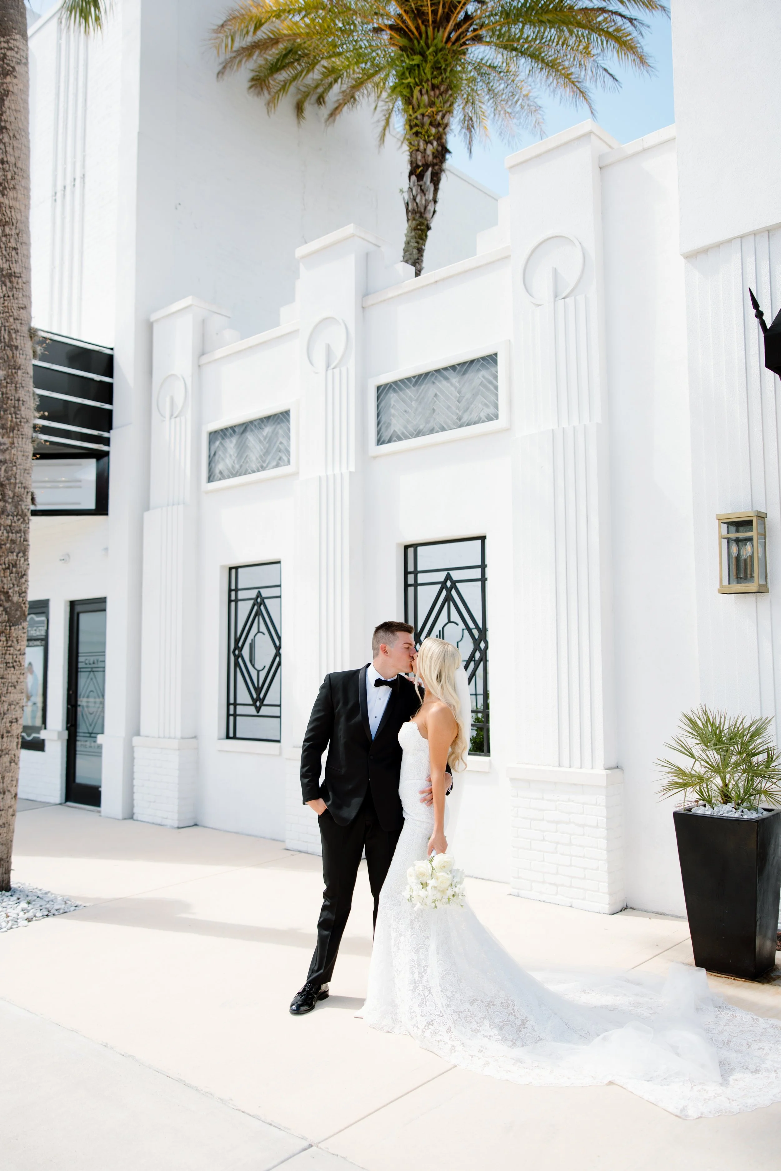 A bride and groom kiss outside a white building with art deco design elements, palm trees, and clear blue sky.
