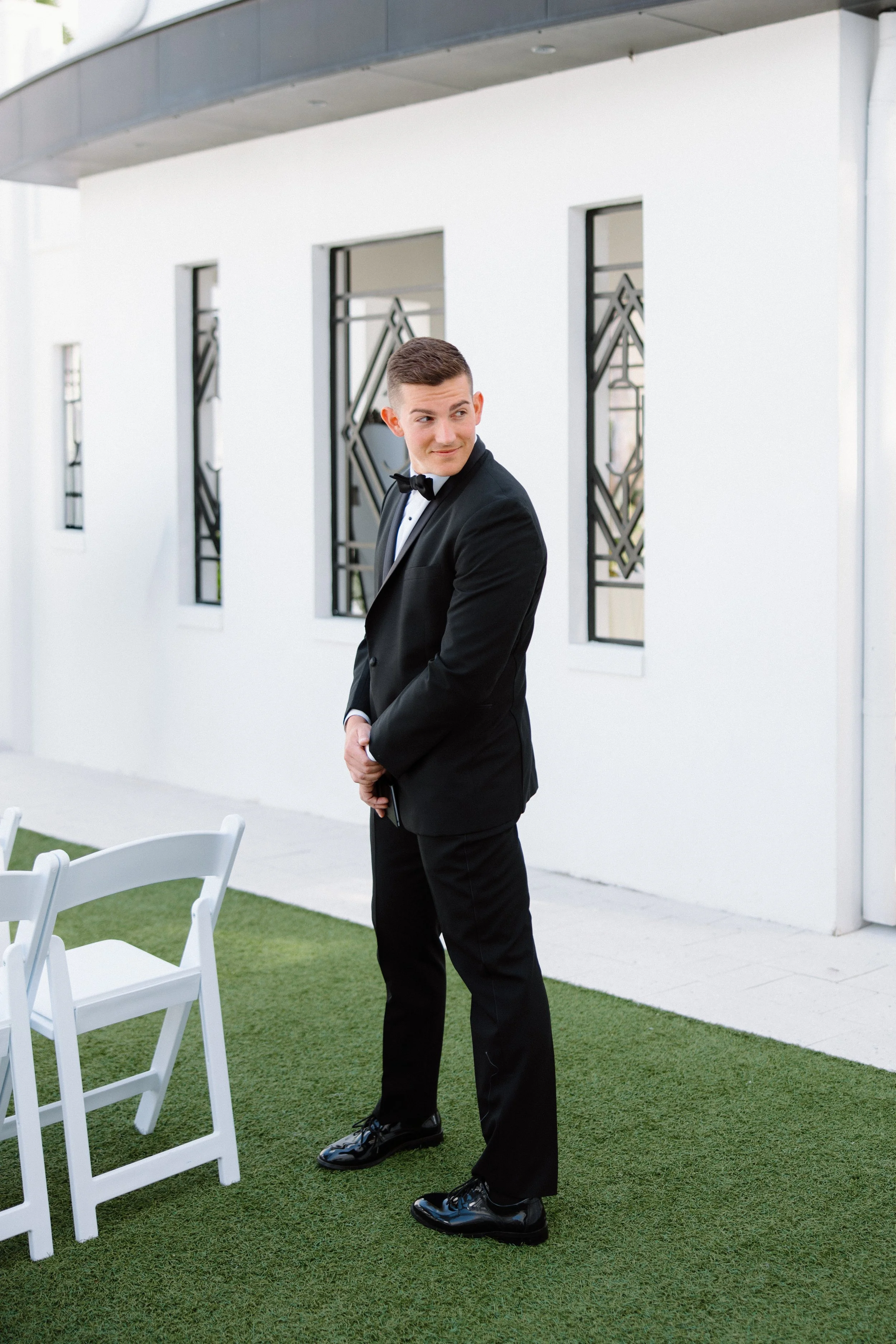 A man wearing a black tuxedo with a bowtie, standing outdoors on a grassy area, near white chairs, with modern white and glass building in the background.