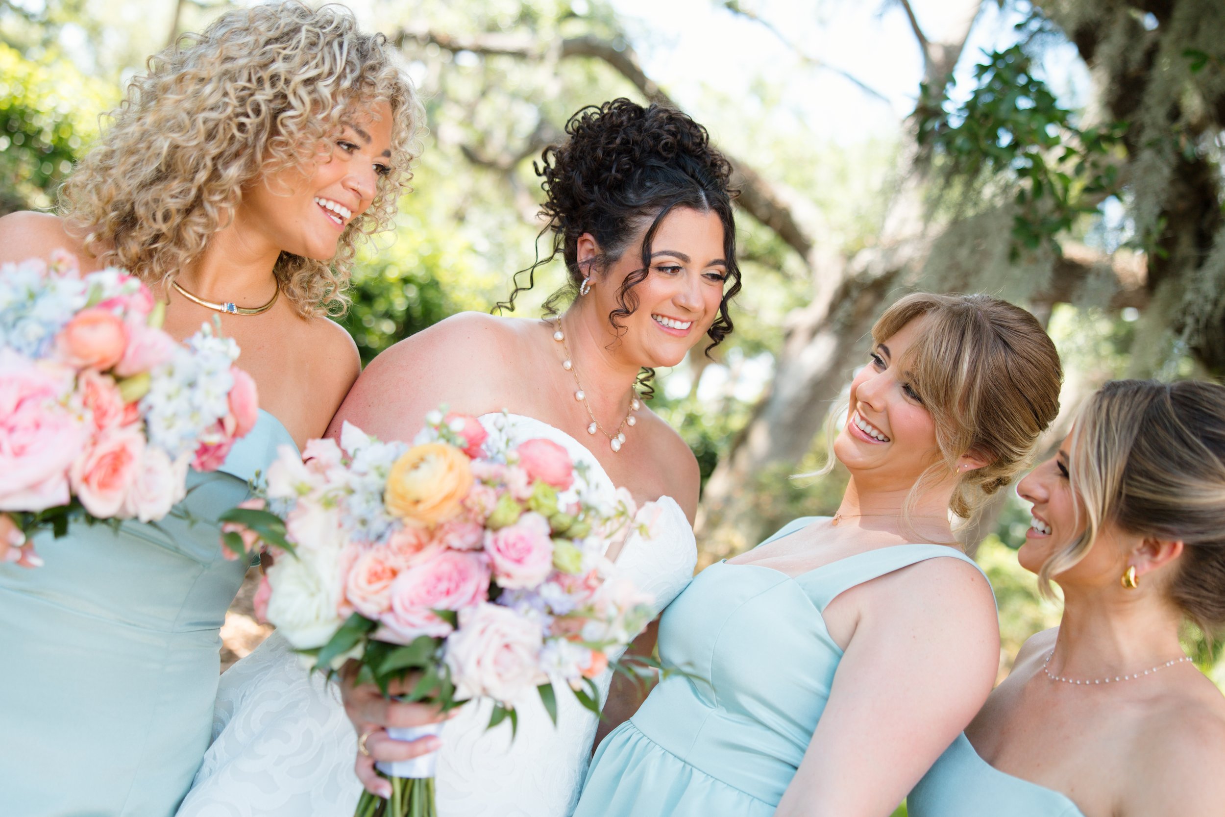 Group of women at a wedding, smiling, with one holding a bouquet of pink and white flowers. They are outdoors with green trees in the background.