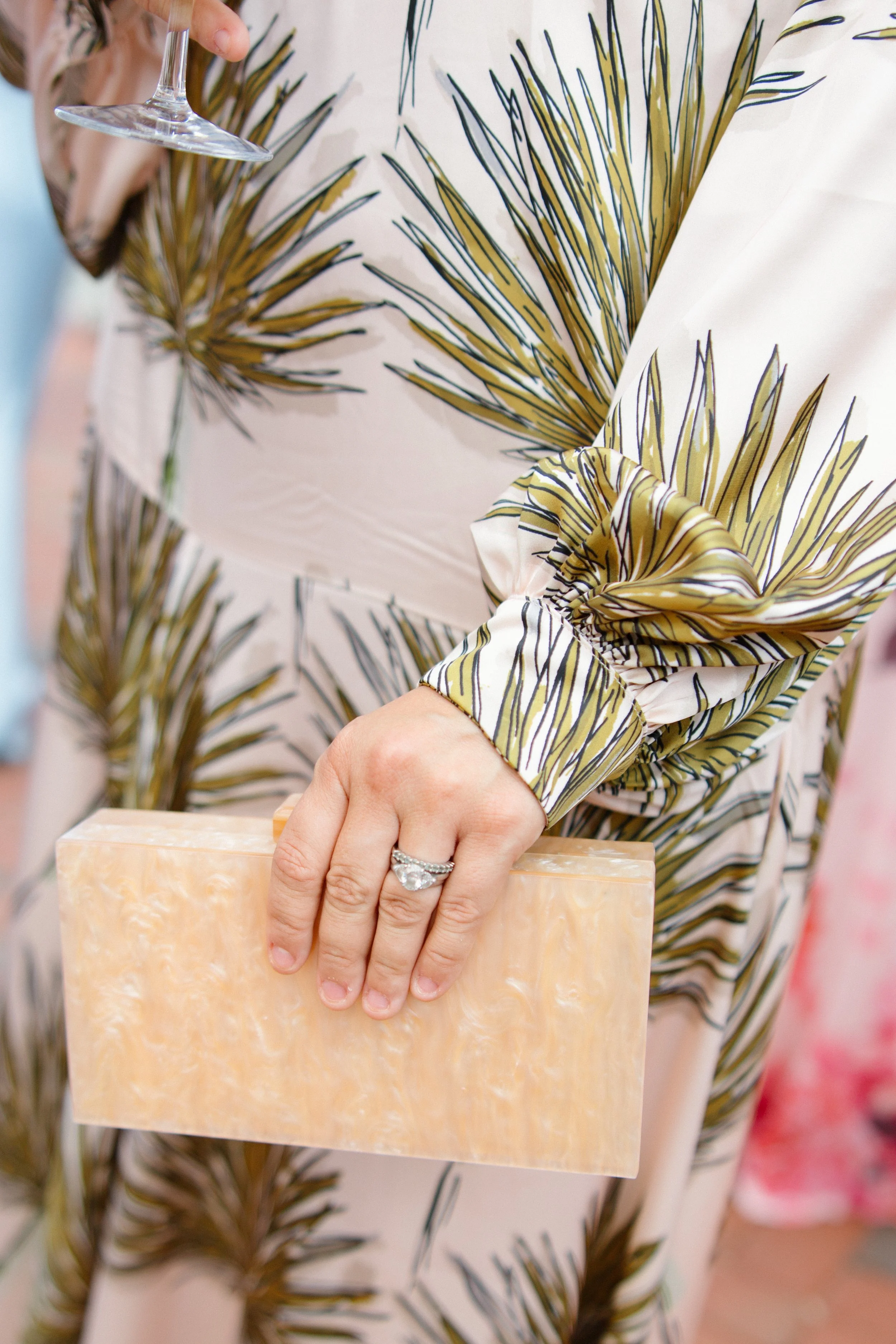 Close-up of a person holding a piece of beige-colored marble with their left hand, which has a large diamond engagement ring. The person's right hand is holding a wine glass. They are wearing a long-sleeved dress with a white background and a brown a