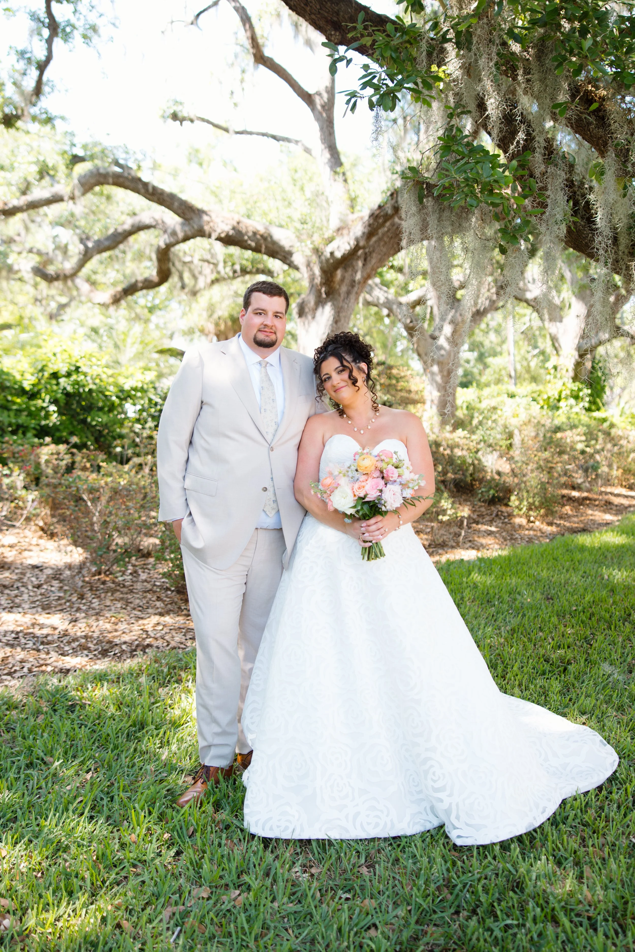 A bride and groom standing together outdoors under a large tree with Spanish moss and lush green foliage. The bride is holding a bouquet of pink and white flowers and is wearing a strapless white wedding gown. The groom is wearing a light-colored sui