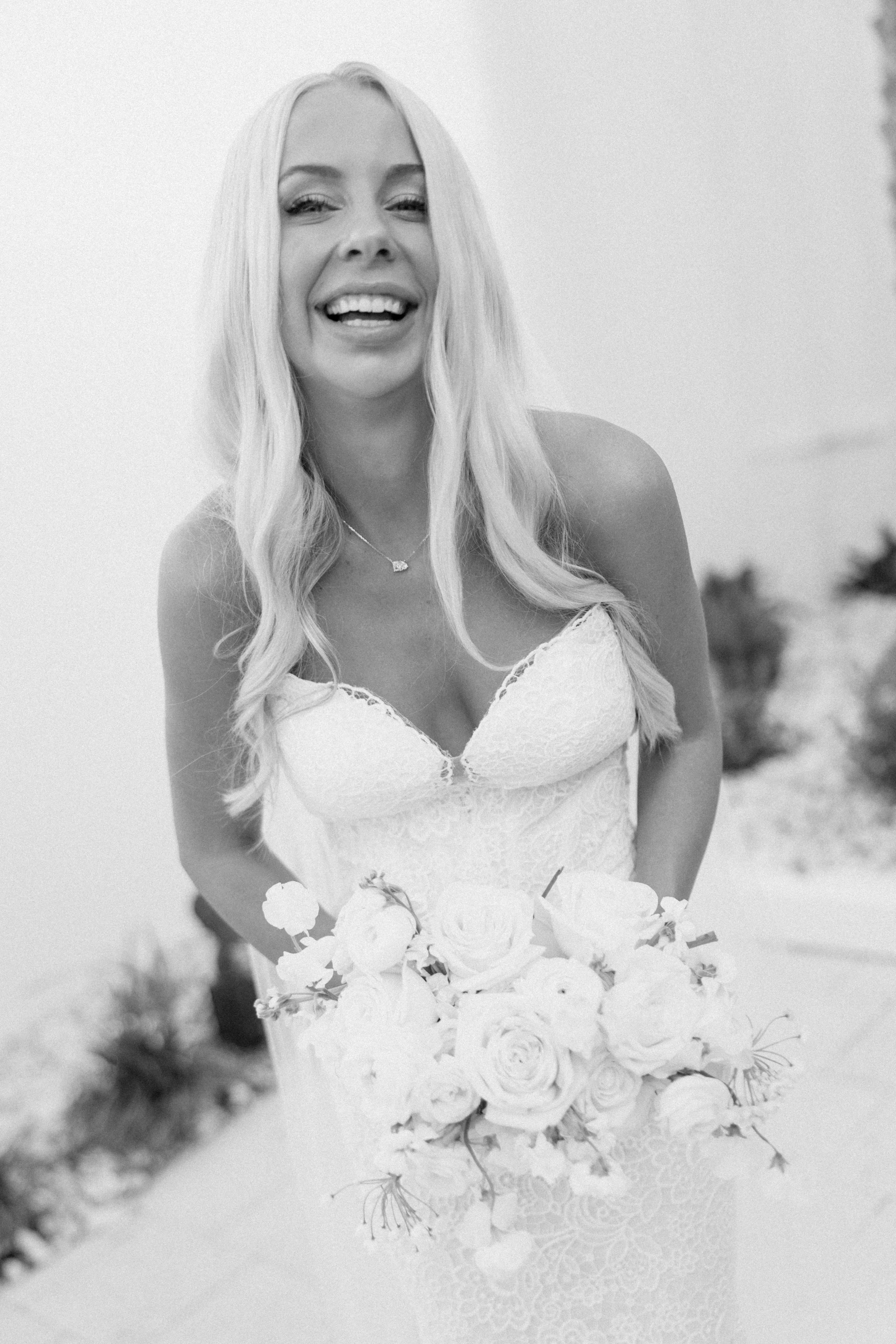 Black and white photo of a woman with long, wavy hair wearing a lace dress, holding a bouquet of roses, smiling, outdoors with blurry background.