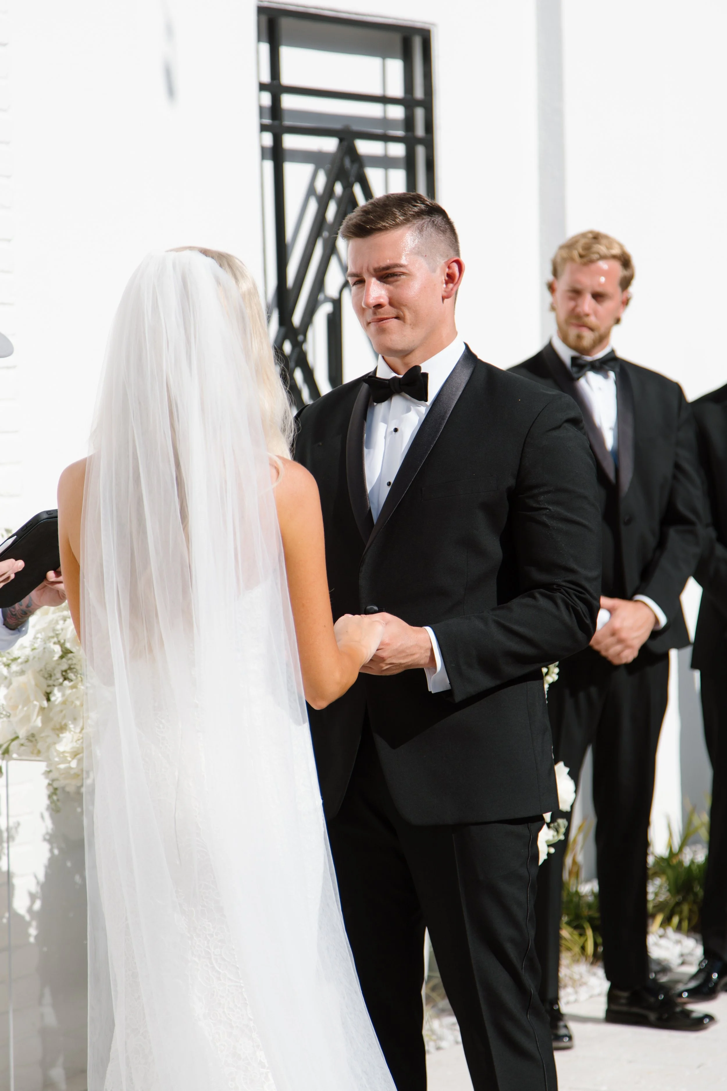 A groom in a black tuxedo and bow tie holding hands with a bride in a white wedding dress and veil during their wedding ceremony outside.