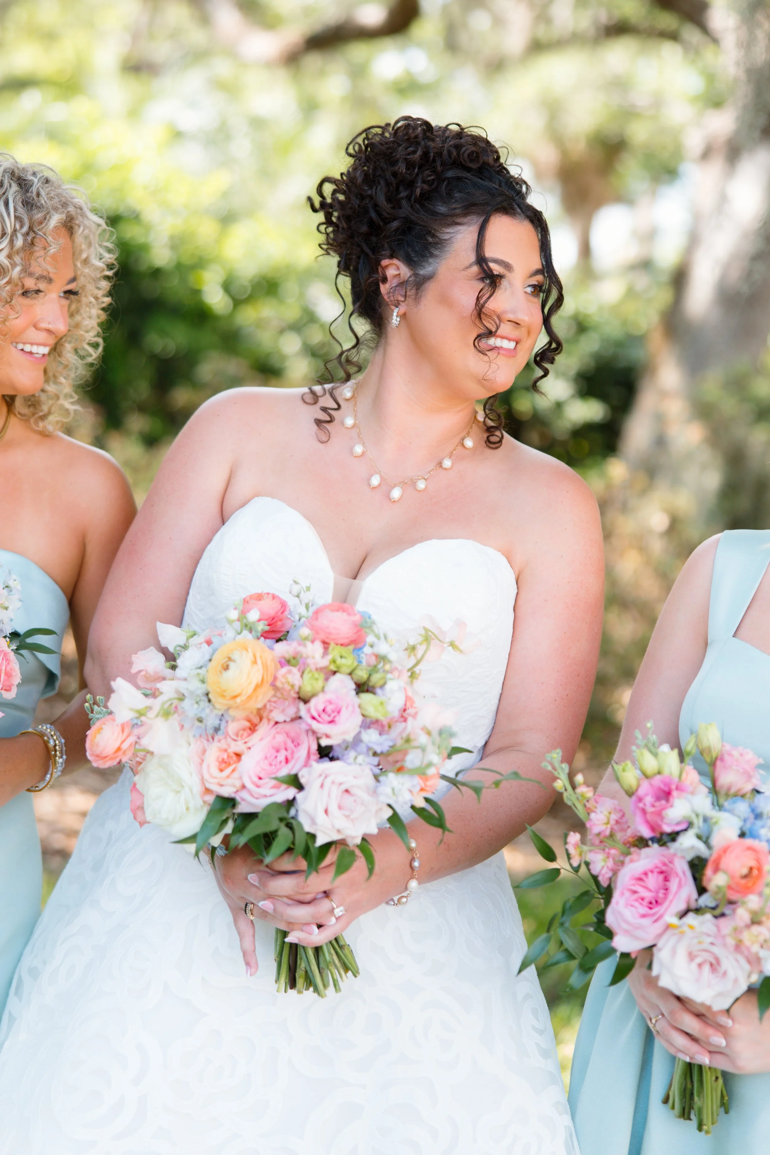A bride in a white wedding dress holding a colorful bouquet of flowers, standing outdoors during daytime, with bridesmaids holding similar bouquets visible on either side.