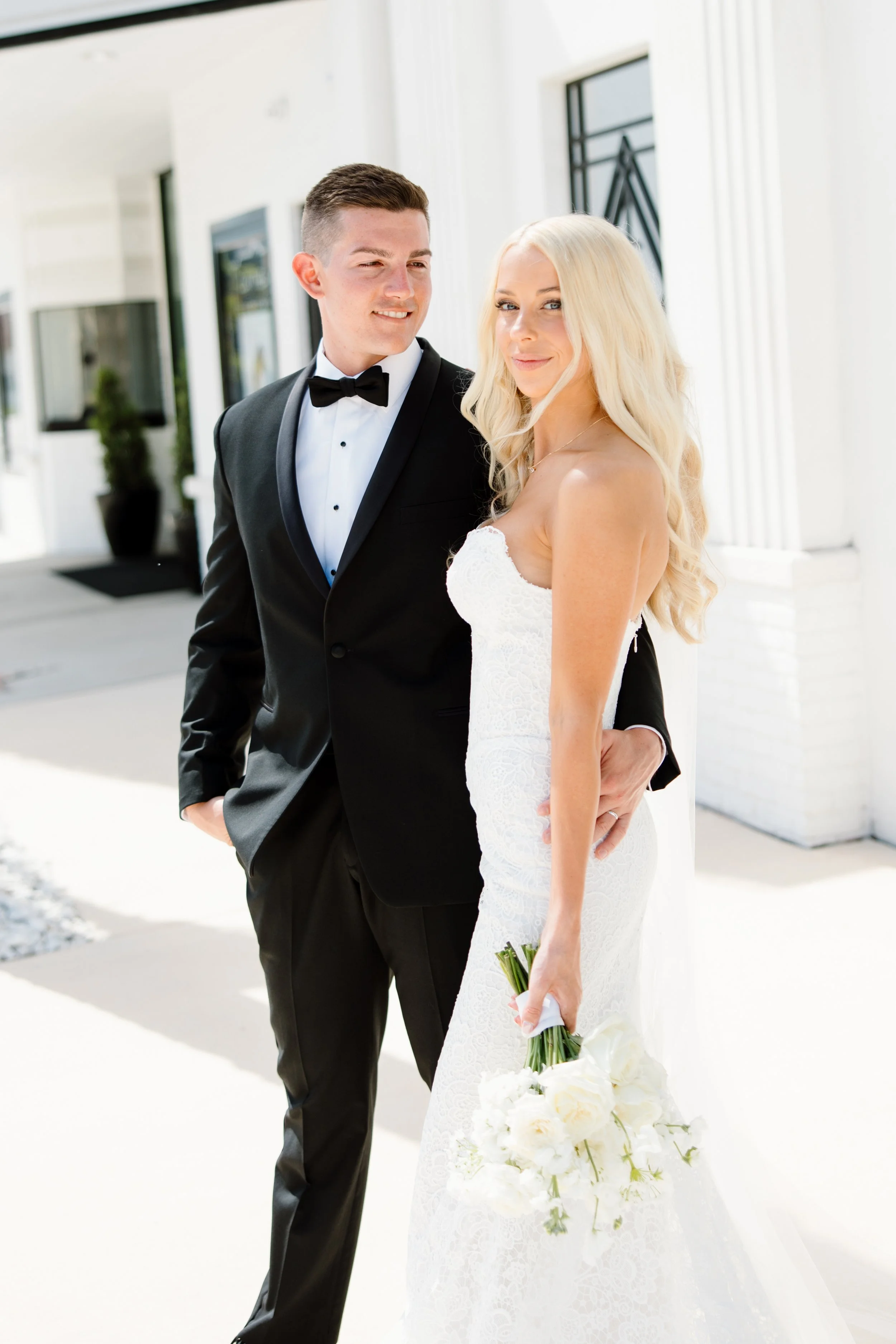 A groom in a black tuxedo with a bow tie standing beside a bride in a white lace wedding dress holding a bouquet of white flowers outside a white building with black window frames.