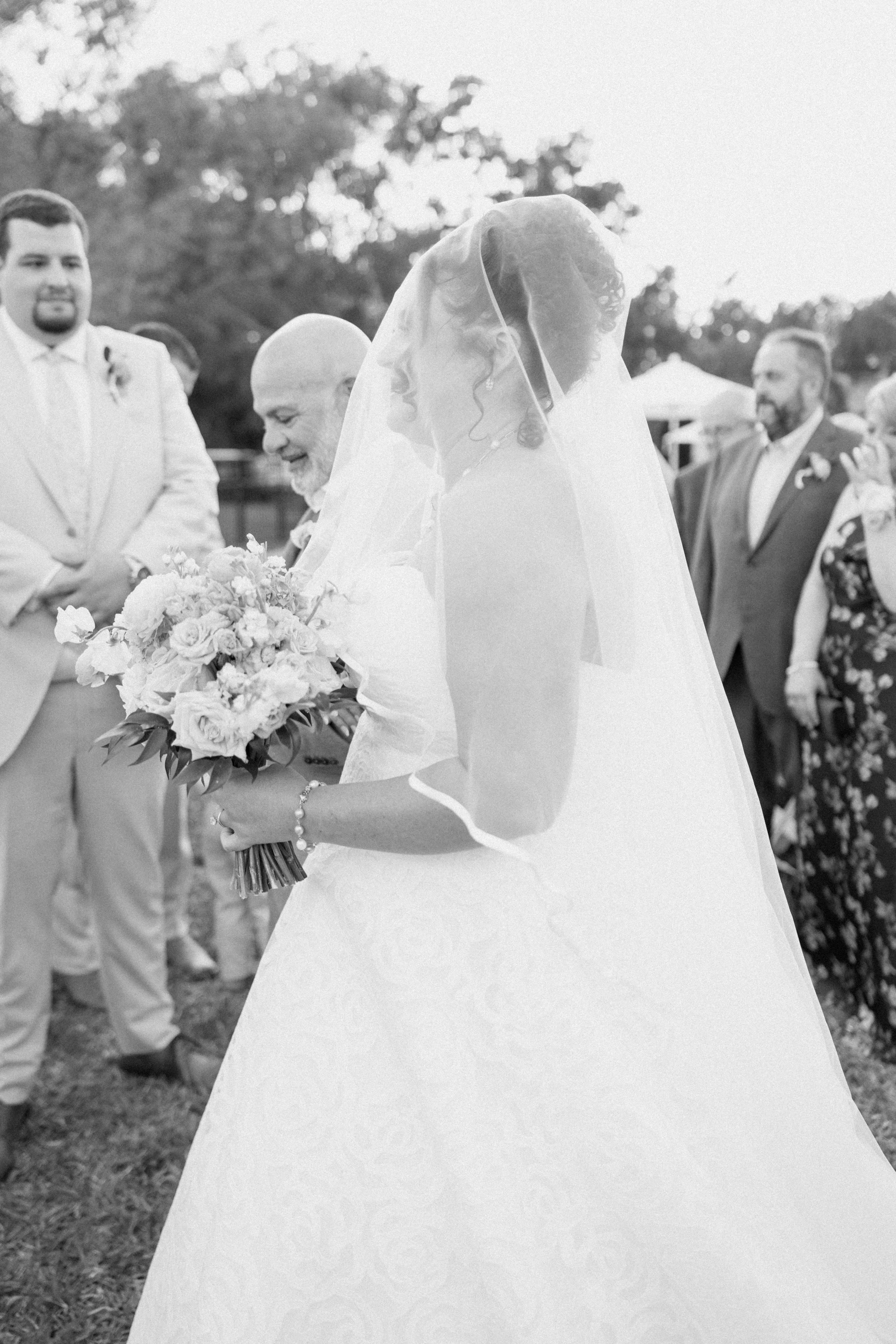 A bride in a white wedding gown and veil holding a bouquet of flowers, surrounded by wedding guests outdoors during daytime.