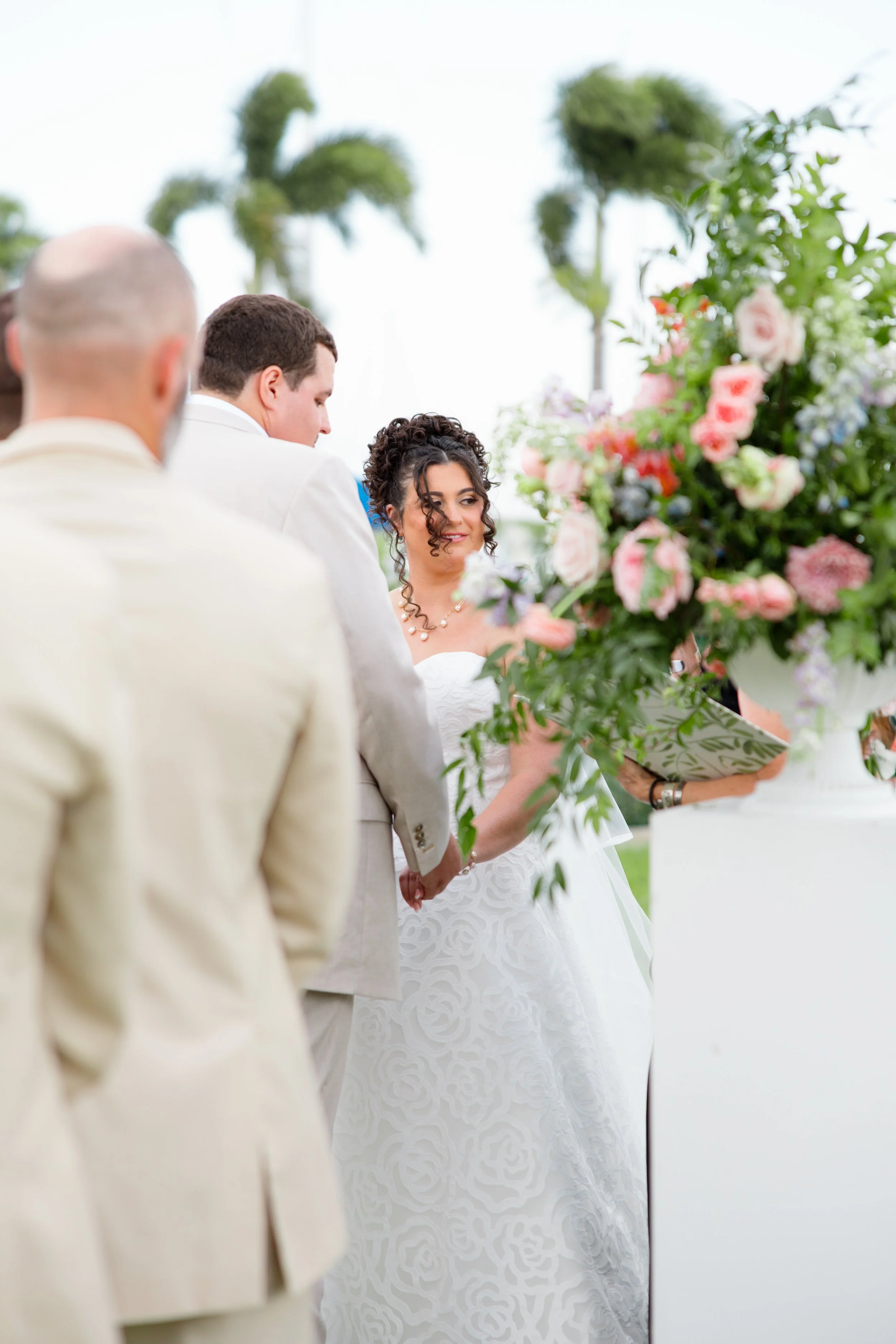 A bride and groom holding hands during a wedding ceremony outside, with wedding officiant and floral arrangements nearby, and palm trees in the background.