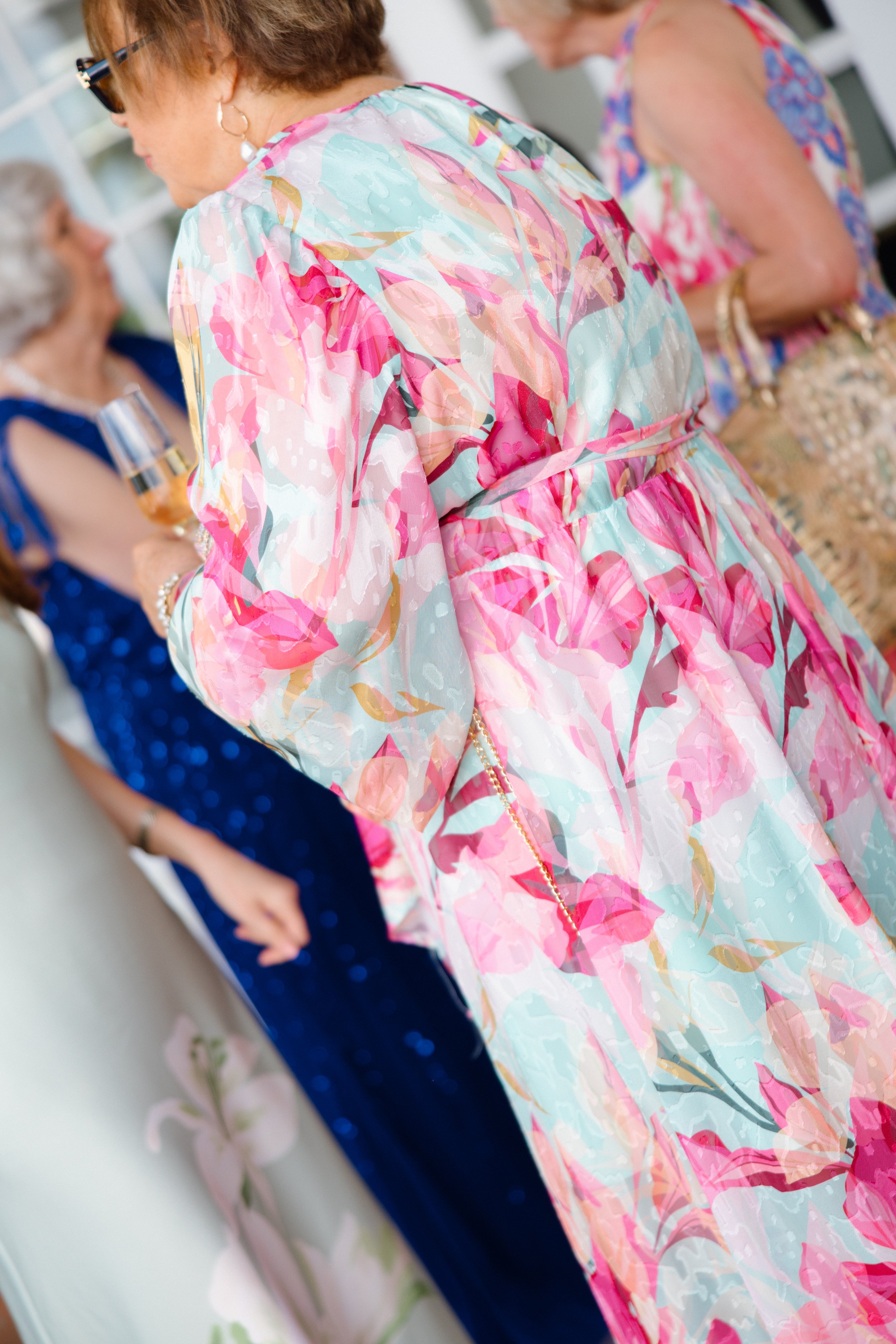The image shows three women dressed in colorful, floral, and elegant dresses, standing close to each other. The focus is on the back of the women, with details of their dresses and accessories visible. The background is blurred.